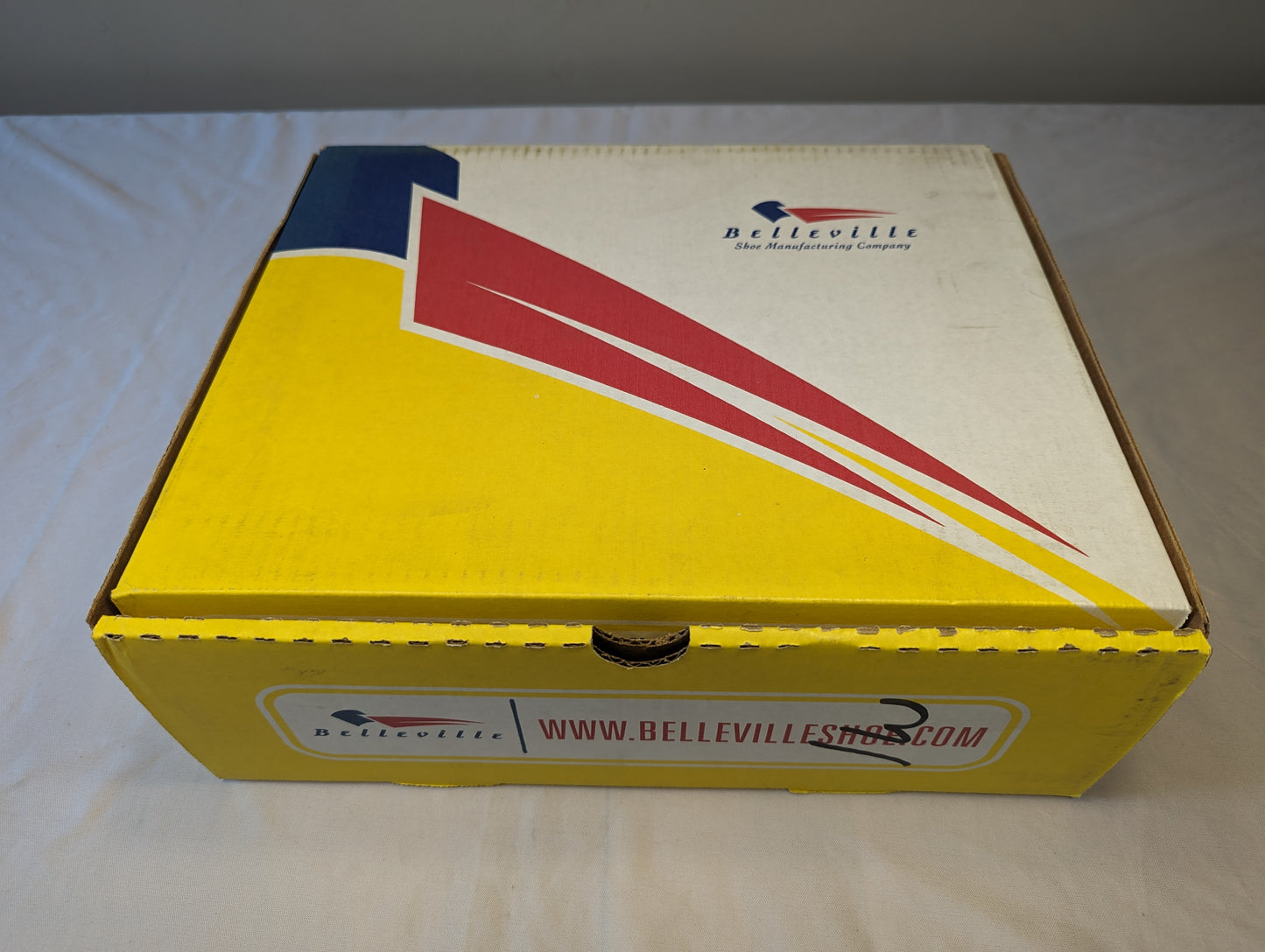A closed cardboard Belleville military boot box, featuring a bold yellow and navy blue design with a red diagonal stripe and the 'BELLEVILLE Shoe Manufacturing Company' logo.