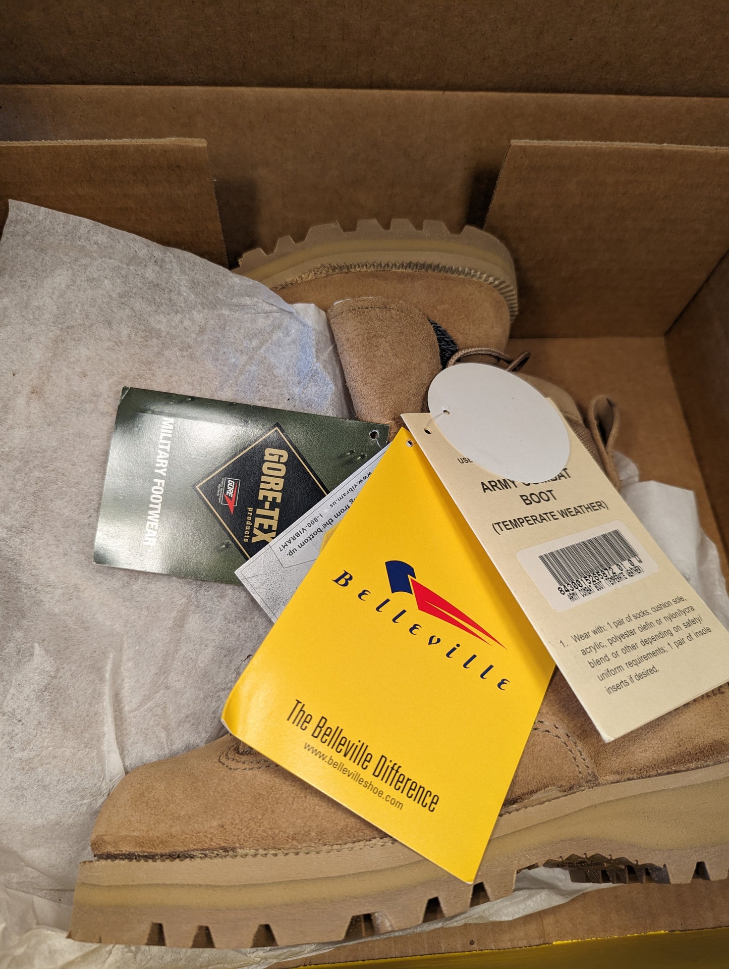 An overhead view into an open cardboard box, showing a new tan Belleville Cold Weather Combat Boot surrounded by tissue paper, with the yellow Belleville brand card and Gore-Tex tag visible.