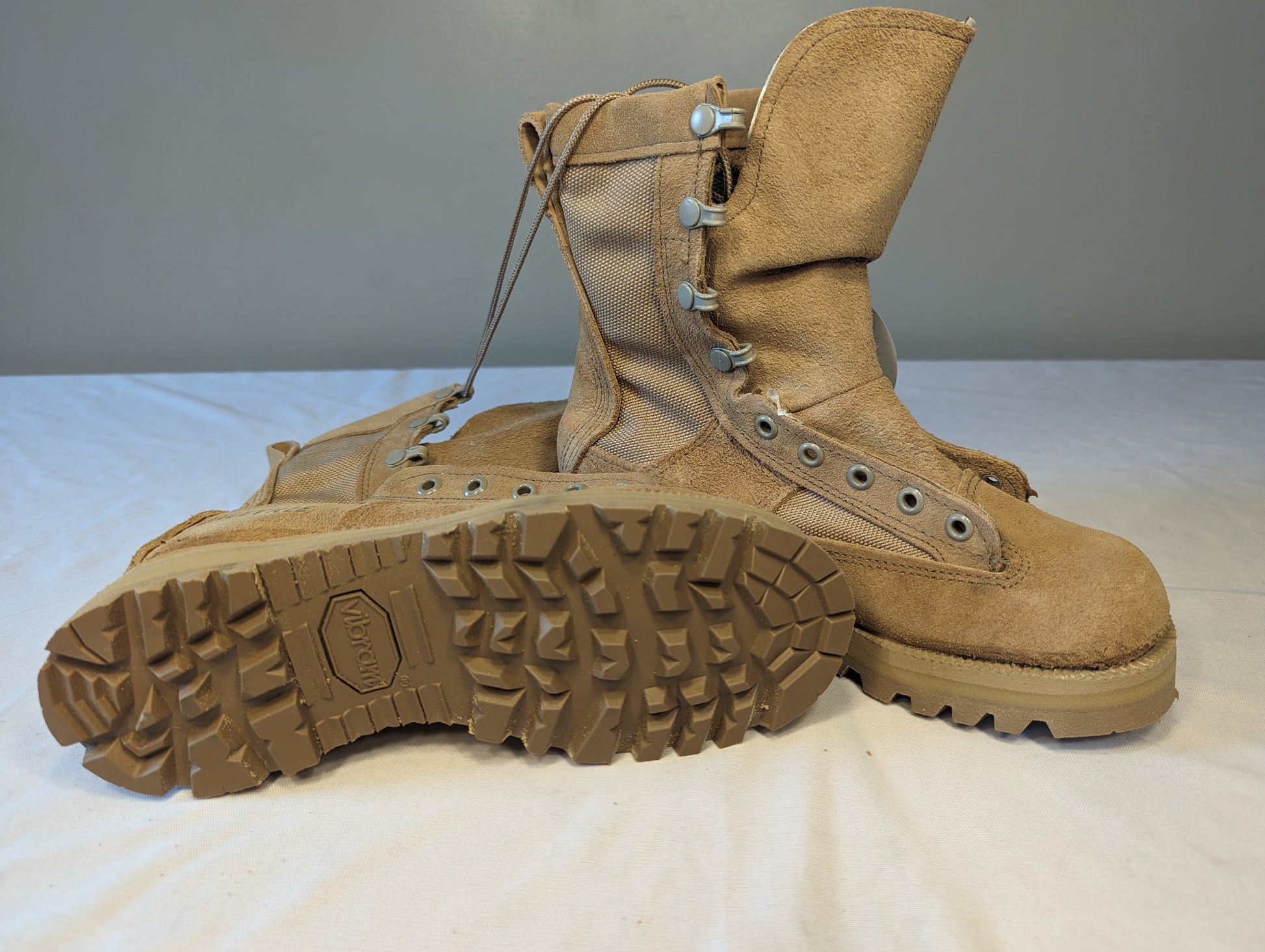 A pair of tan Belleville Cold Weather Combat Boots on a white background, with one boot lying on its side to display the aggressive Vibram sole tread pattern and the other standing upright.