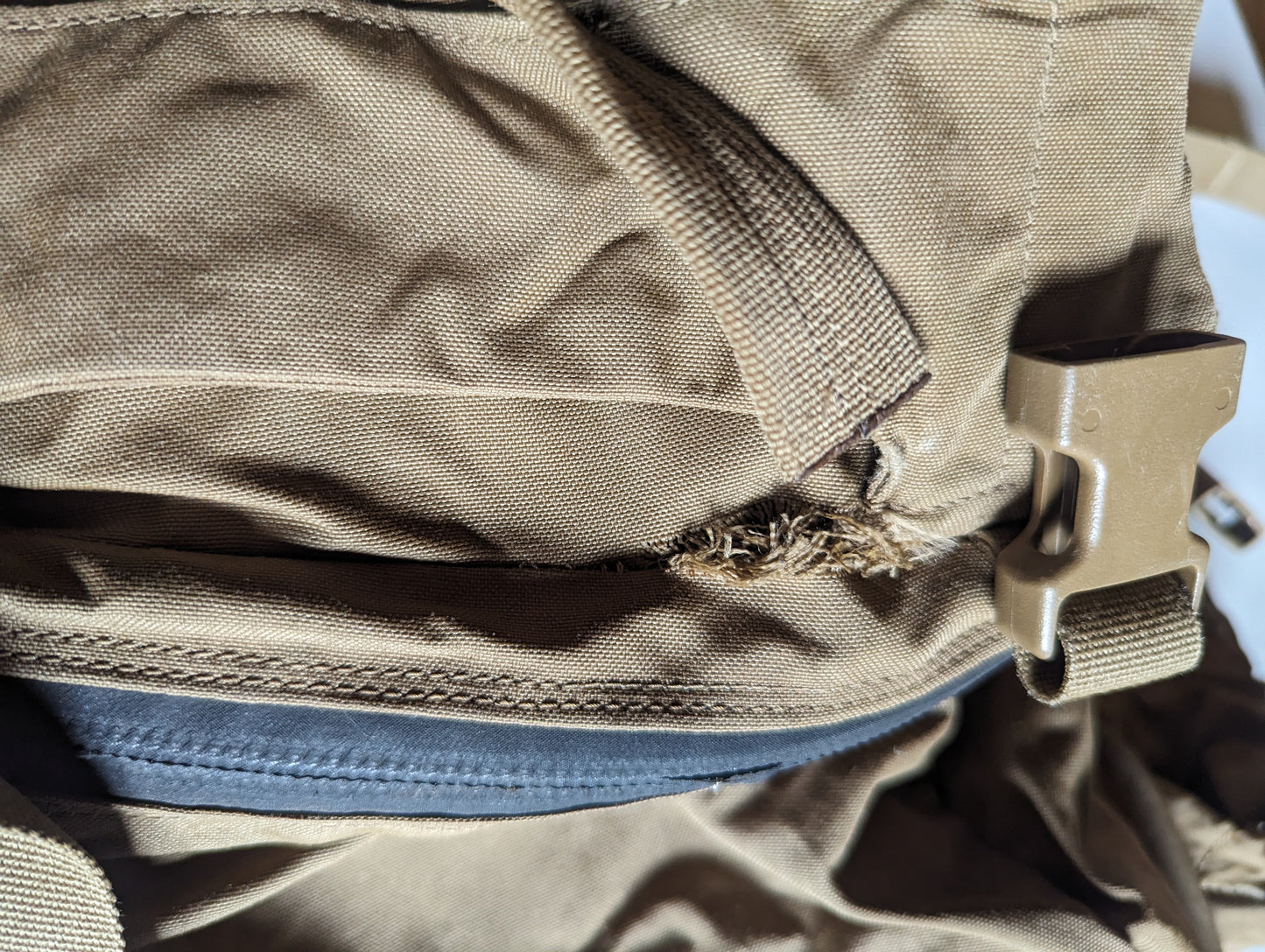 An extreme close-up of a Coyote Brown tactical pack, highlighting a large tear and severe fraying in the nylon fabric where the webbing strap and plastic buckle meet the zipper line, documenting surplus damage.