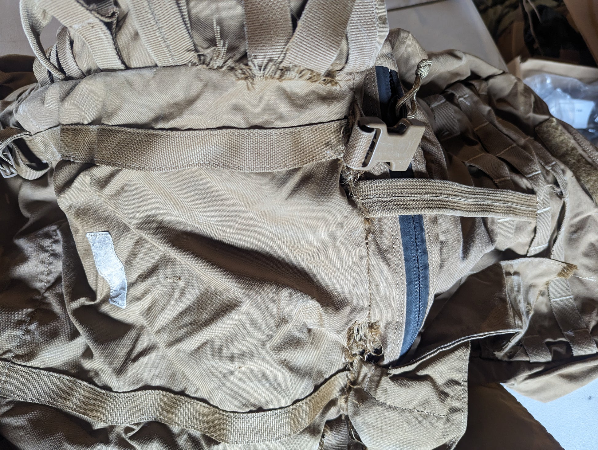 A close-up photograph of a Coyote Brown tactical pack, highlighting severe fraying and tearing damage to the nylon fabric near a broken black zipper and plastic quick-release buckle.