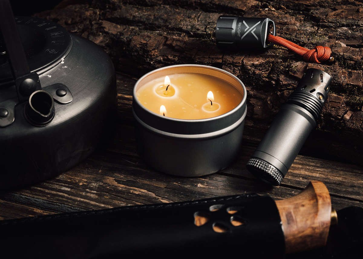 A close-up of an open candleTIN with three lit wicks, surrounded by rugged camping gear including a kettle and a fire starter on a dark, rustic wooden surface.