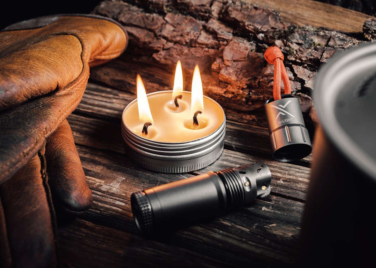 A close-up of an open candleTIN with three lit wicks, resting on a dark wooden surface next to a smaller fire starter and a gloved hand, surrounded by camping gear.
