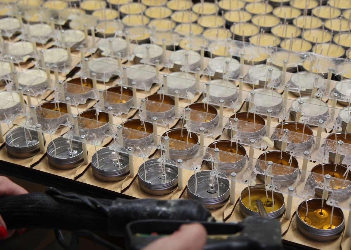 A close-up view of the candleTIN production process, showing rows of metal tins being filled with yellow wax, with wicks held upright by a wooden jig and clear plastic sheets.