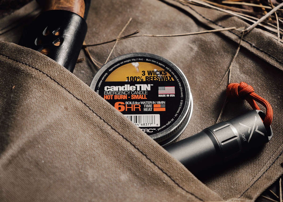 A close-up of a CANDLE TIN (3 WICKS, 100% BEESWAX) tucked into a canvas pocket alongside a fire starter, emphasizing its compact size and features.