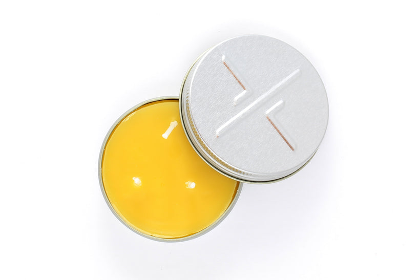 An overhead studio shot of a small, open candleTIN revealing the smooth yellow beeswax and two short wicks, with the embossed lid resting next to the tin on a white background.