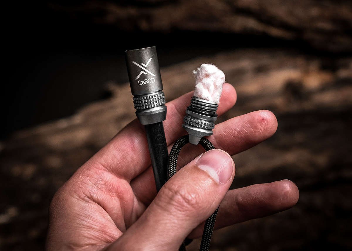 A close-up of a person's hand holding a gunmetal fireROD fire starter and a small bundle of white tinder, against a dark, heavily textured wooden background.
