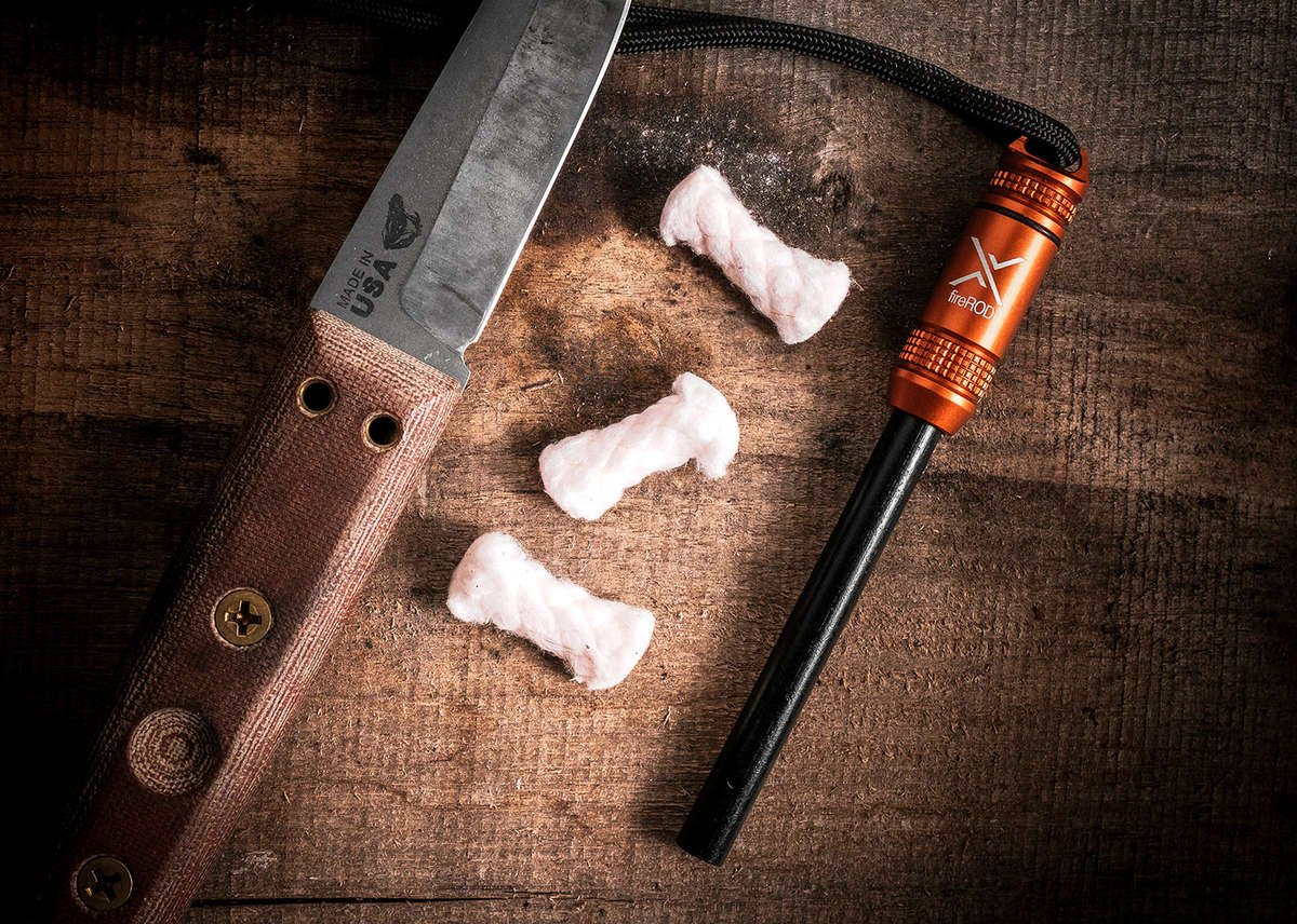 A close-up of a fixed-blade knife with a tan handle and an orange fireROD fire starter, resting on a rustic wooden surface next to three bundles of emergency tinder.