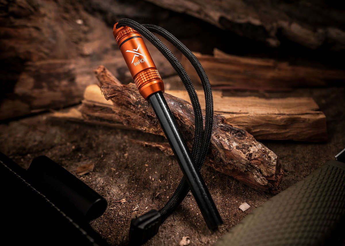 A close-up of the fireROD fire starter lying on dark, chopped kindling, featuring an orange anodized aluminum handle and a black ferrocerium rod with a black paracord lanyard.