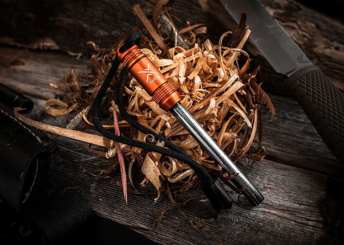 A close-up of the fireROD fire starter with an orange anodized aluminum handle, resting on a large pile of wood shavings/tinder, with the tip of a large camping knife visible in the background.