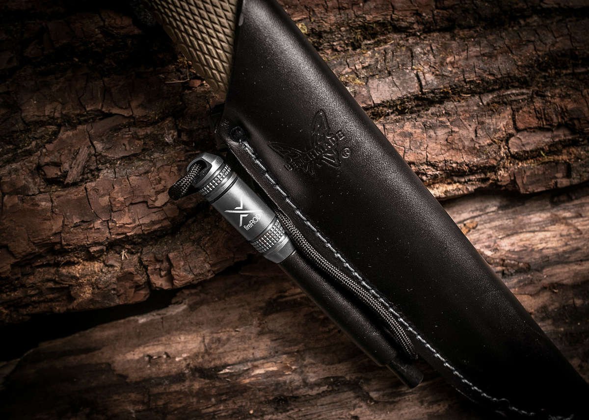 A close-up of a silver fireROD fire starter secured in the accessory loop of a black leather knife sheath resting on a dark, heavily textured wooden surface.