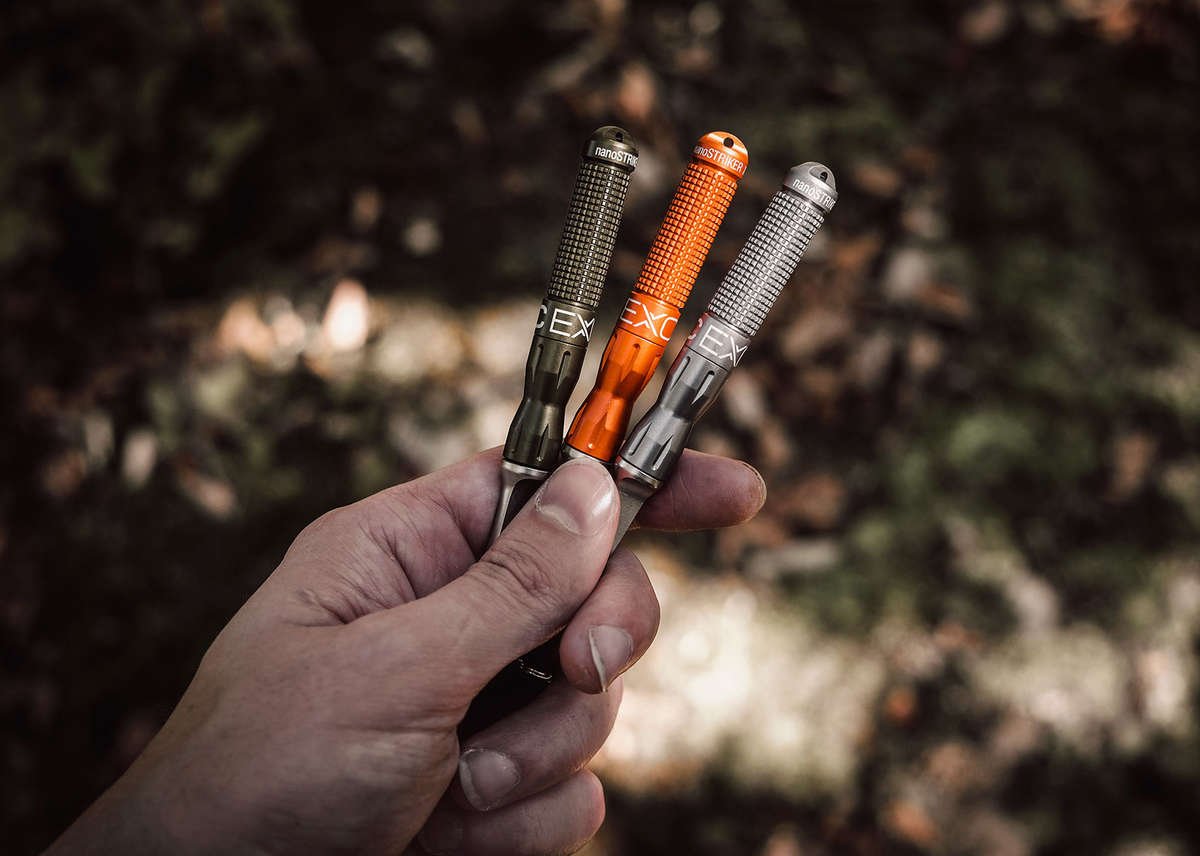 A close-up of a hand holding three nanoSTRIKER XL fire starters—gunmetal, olive green, and orange—against a blurred, dark natural background, emphasizing their compact size.