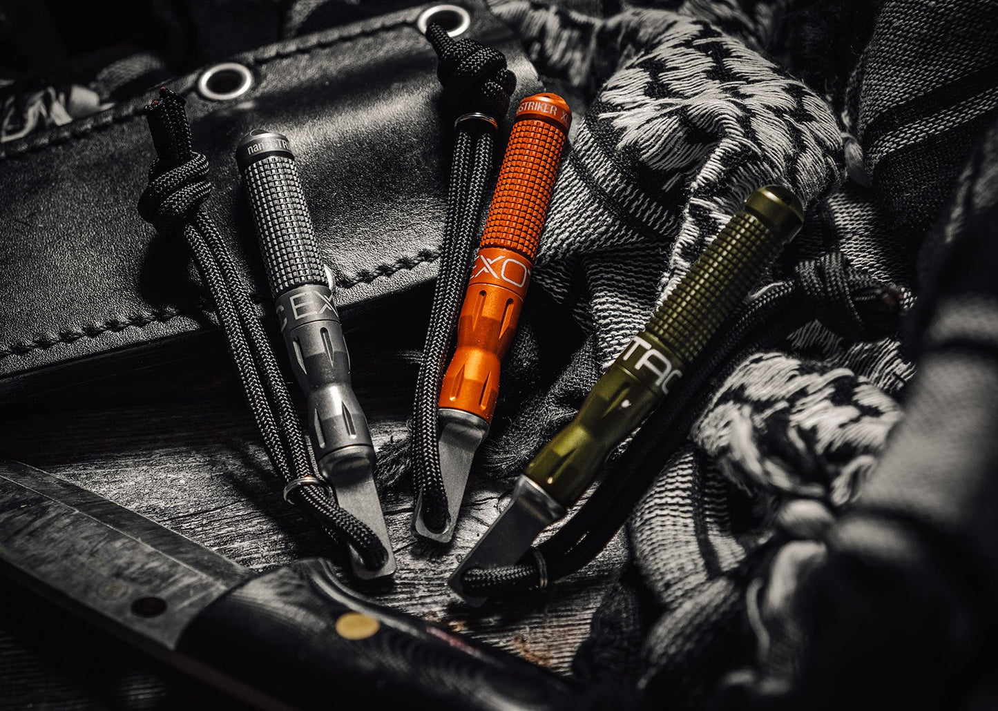 A close-up of three nanoSTRIKER XL fire starters (black, orange, and olive green) resting on a dark, wooden surface among tangled paracord, a tactical knife, and a patterned scarf.