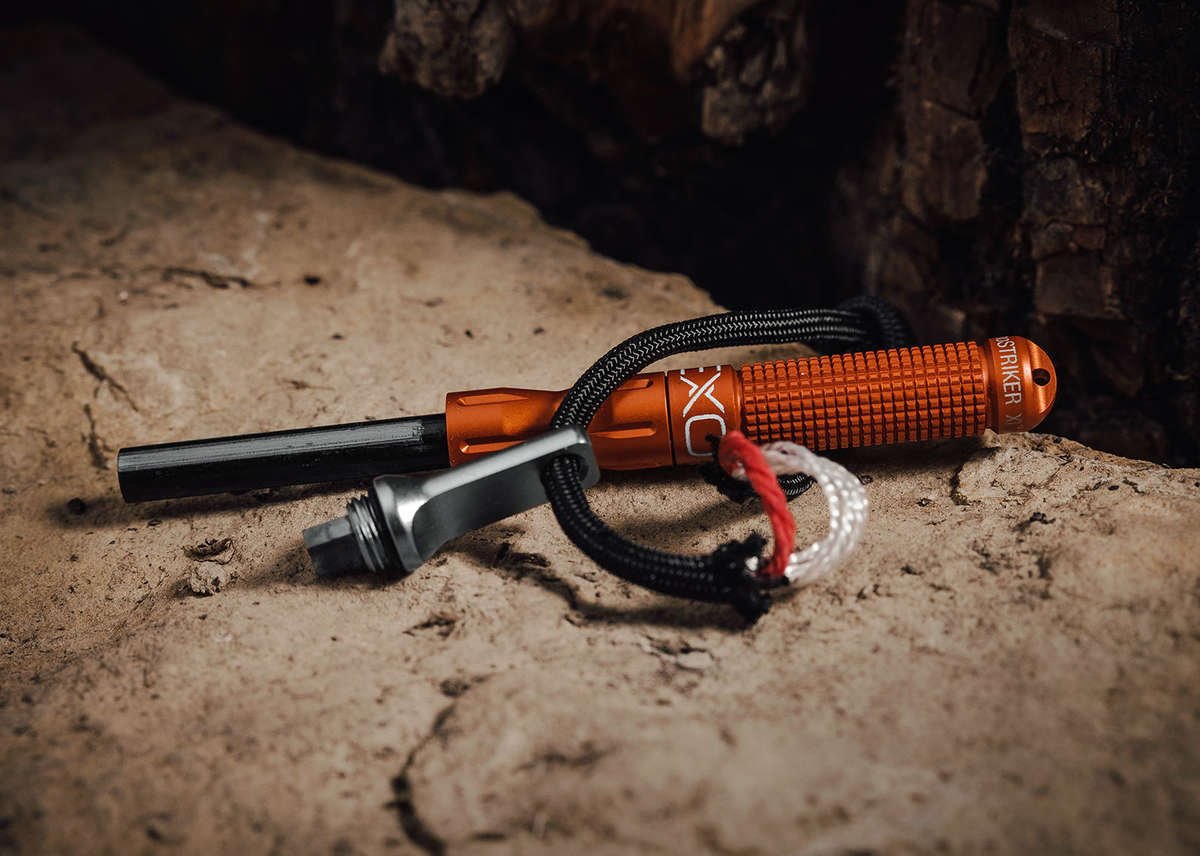 A close-up of the orange nanoSTRIKER XL fire starter disassembled, showing the knurled aluminum handle, the black ferrocerium rod, and the silver striker, resting on a rough, textured rock surface.