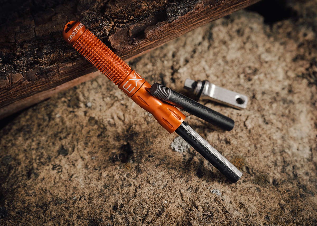 A close-up of the orange nanoSTRIKER XL fire starter disassembled, showing the knurled aluminum handle, the black ferrocerium rod, and the silver striker, resting on a rough rock and wood background.
