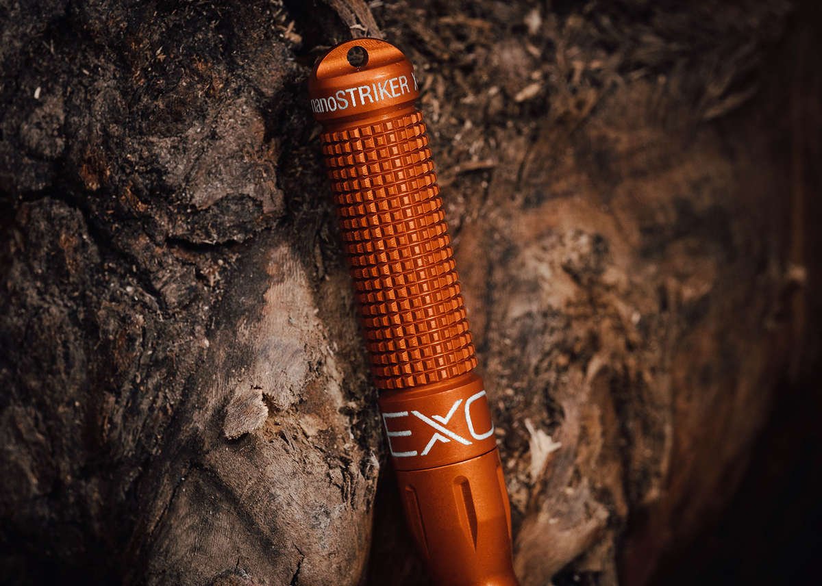 A close-up of a bright orange nanoSTRIKER XL fire starter standing upright in a deep crevice of dark, heavily textured tree bark, emphasizing its rugged construction.