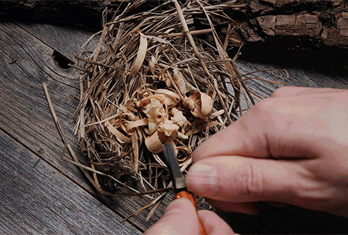 Animated clip showing a person's hand striking the nanoSTRIKER XL to generate a shower of sparks that successfully ignite a bundle of dry tinder resting on dark wood.