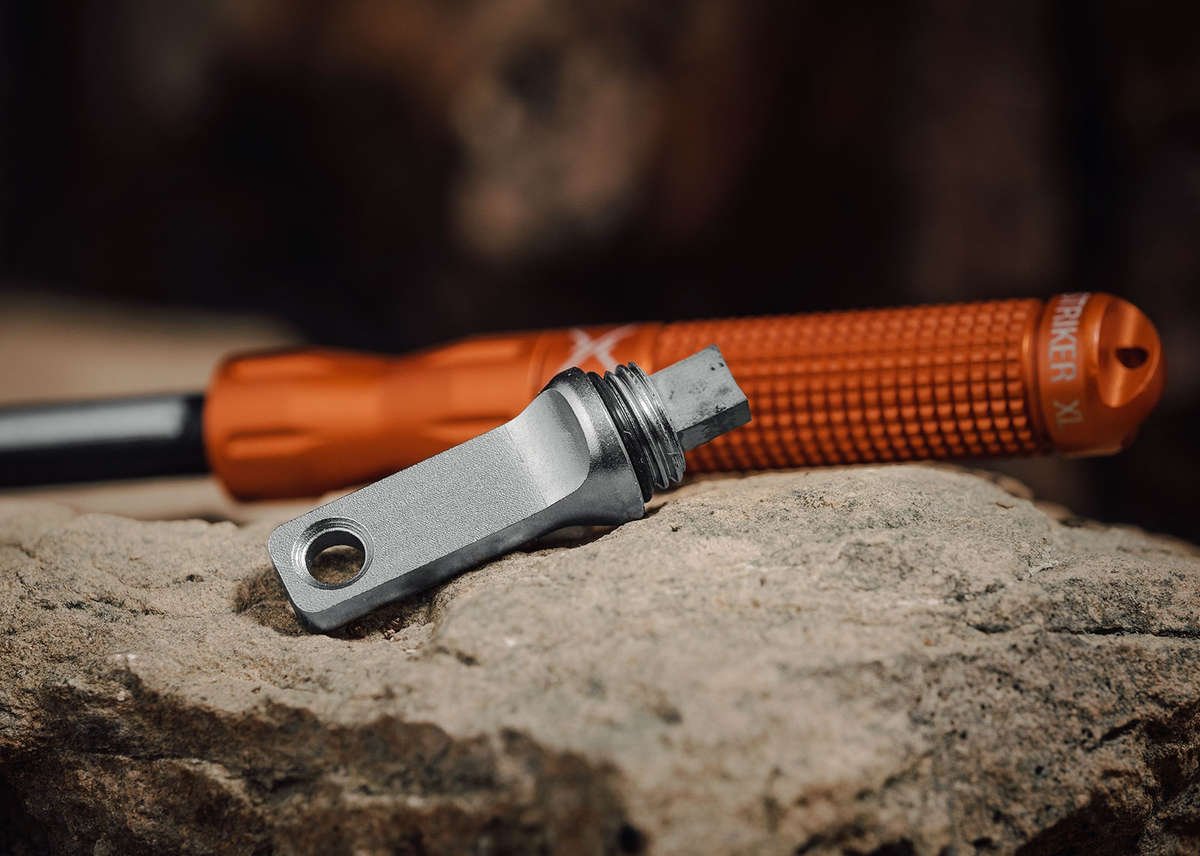 A close-up of the silver metal striker piece from the nanoSTRIKER XL resting on a rough rock surface, with the orange knurled handle blurred in the background.