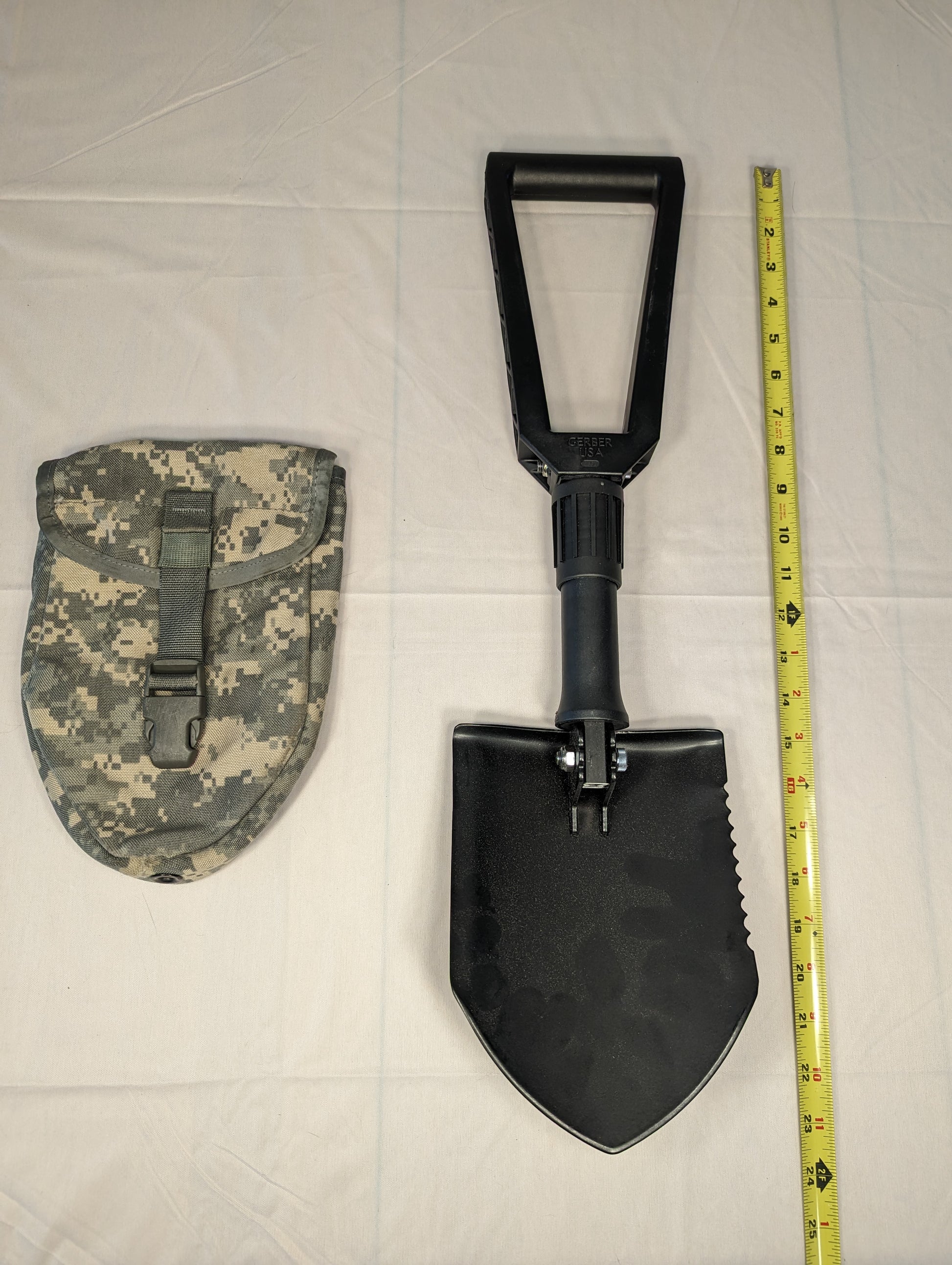 A black Gerber Entrenching Tool shown fully extended next to its ACU digital camouflage carrying pouch, with a yellow tape measure placed alongside to demonstrate the full length.