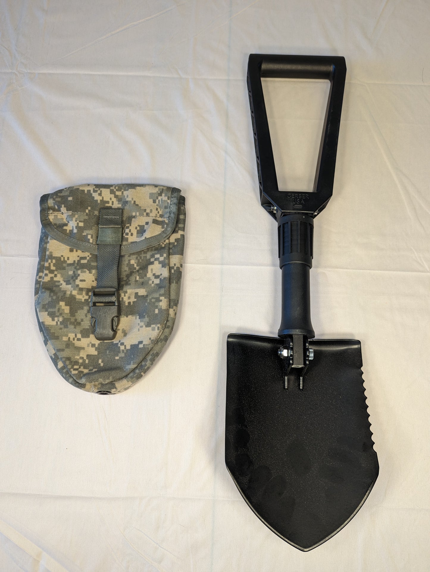 A black Gerber Entrenching Tool shown fully extended, featuring a serrated steel shovel blade and a D-handle, displayed next to its ACU digital camouflage carrying pouch on a white background.