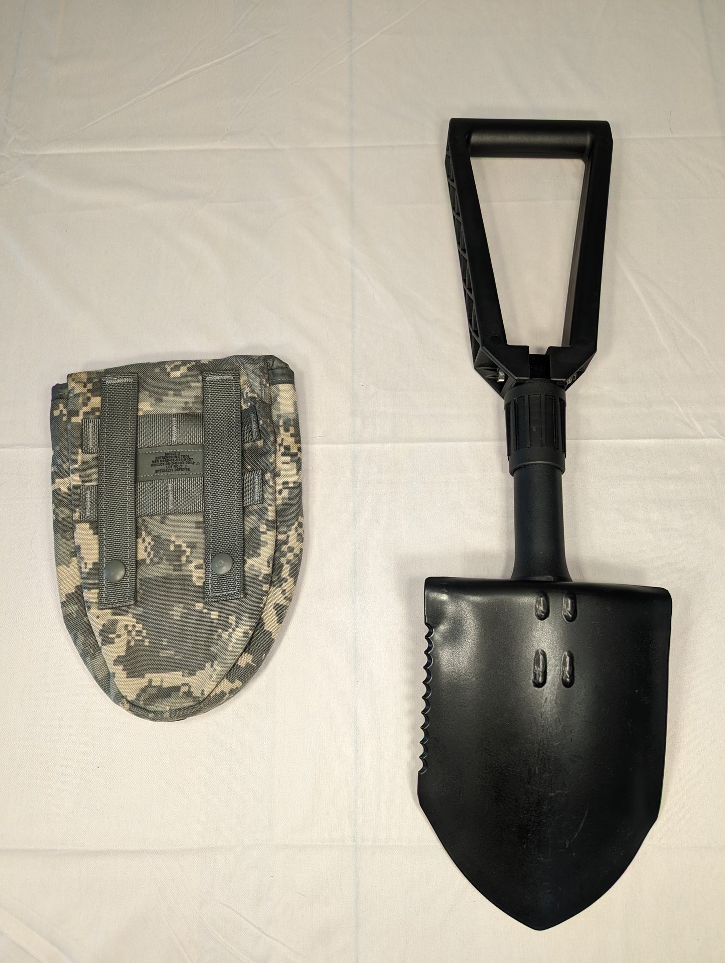 A black Gerber Entrenching Tool shown fully extended next to its ACU digital camouflage carrying pouch, displaying the serrated steel blade and folding handle on a white background.