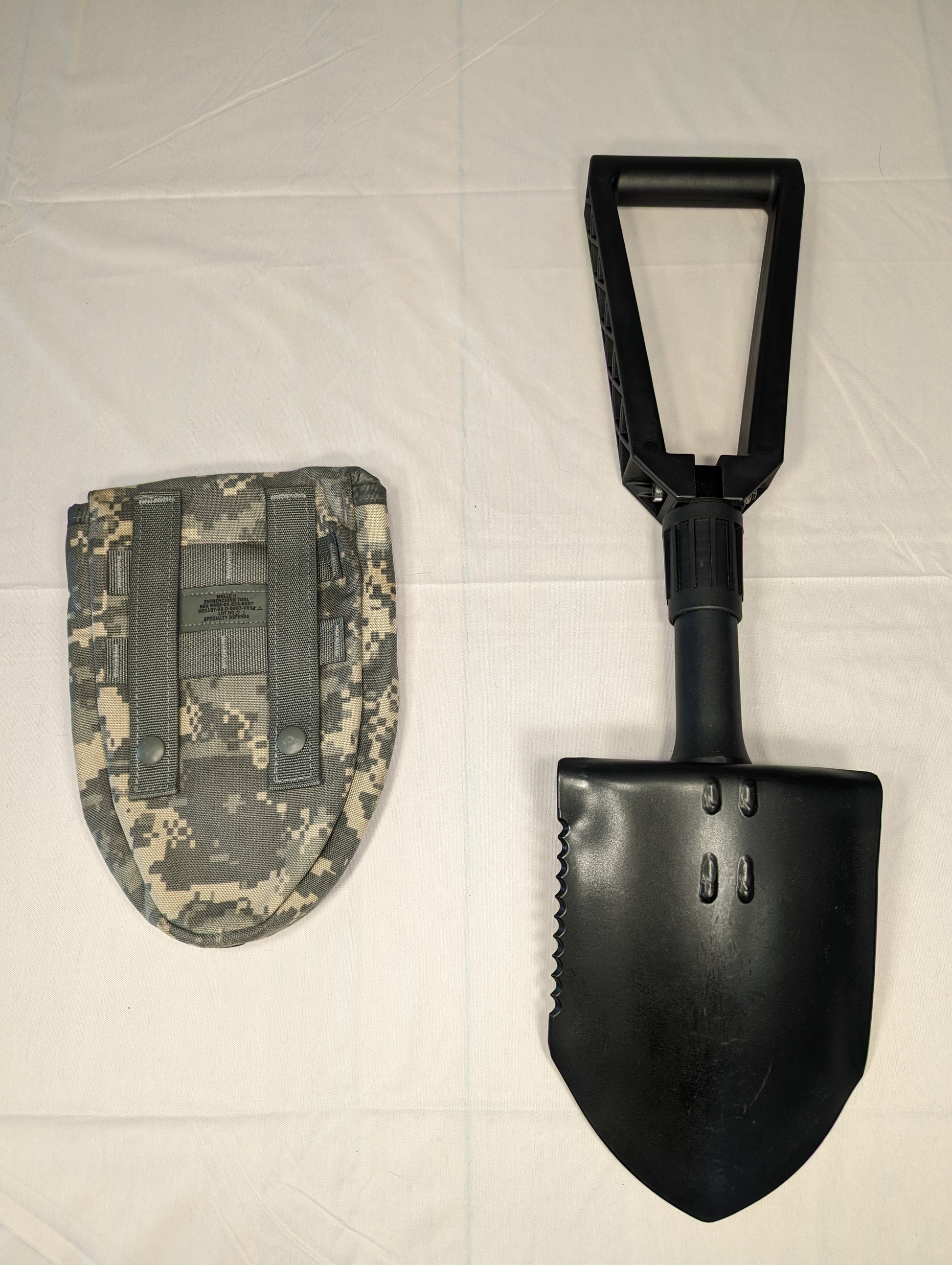 A black Gerber Entrenching Tool shown fully extended next to its ACU digital camouflage carrying pouch, displaying the serrated steel blade and folding handle on a white background.
