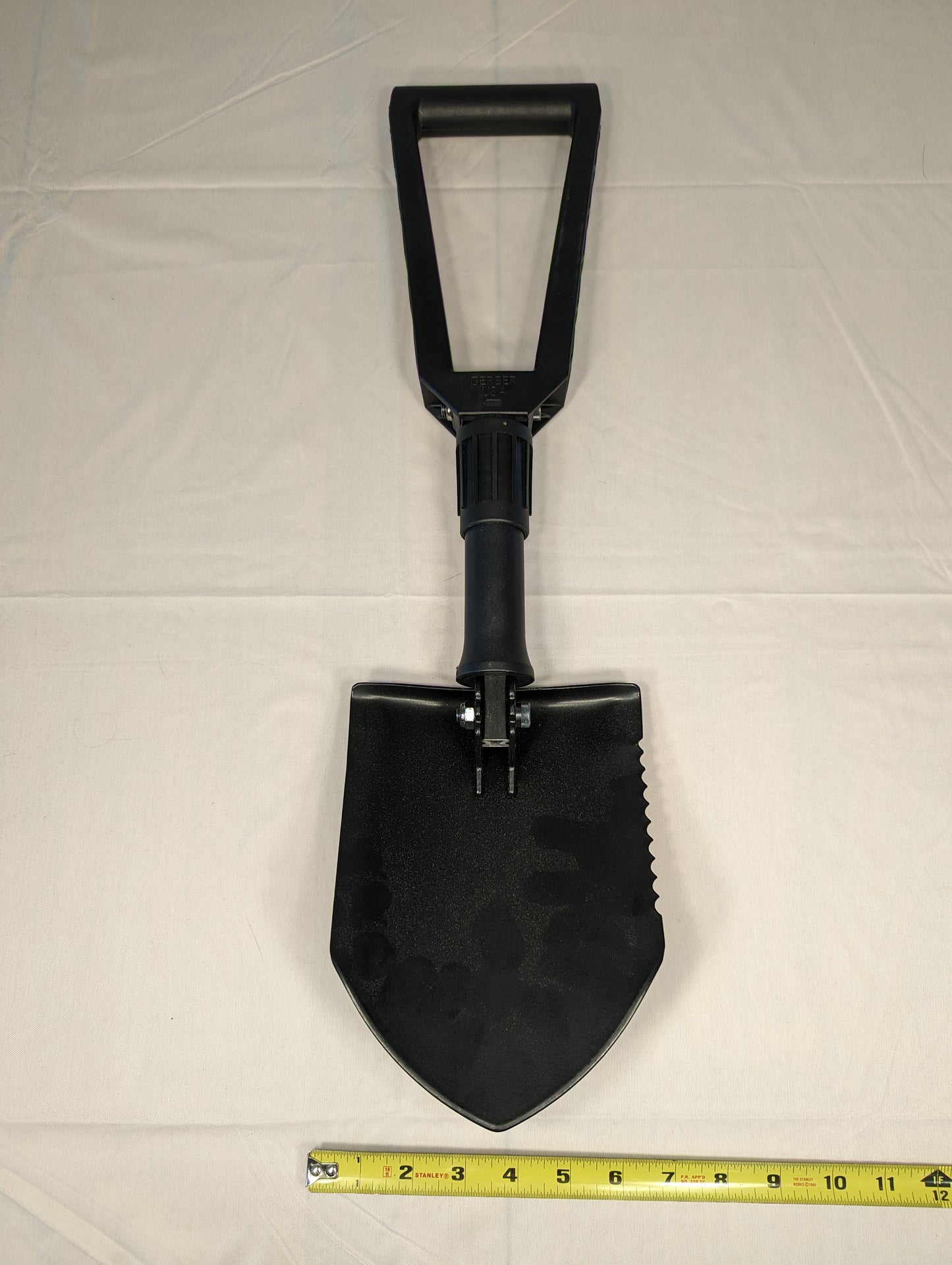 A black Gerber Entrenching Tool (E-Tool) shown fully extended on a white background, featuring a serrated steel shovel blade and a black plastic D-handle, with a yellow tape measure placed at the bottom to demonstrate scale.