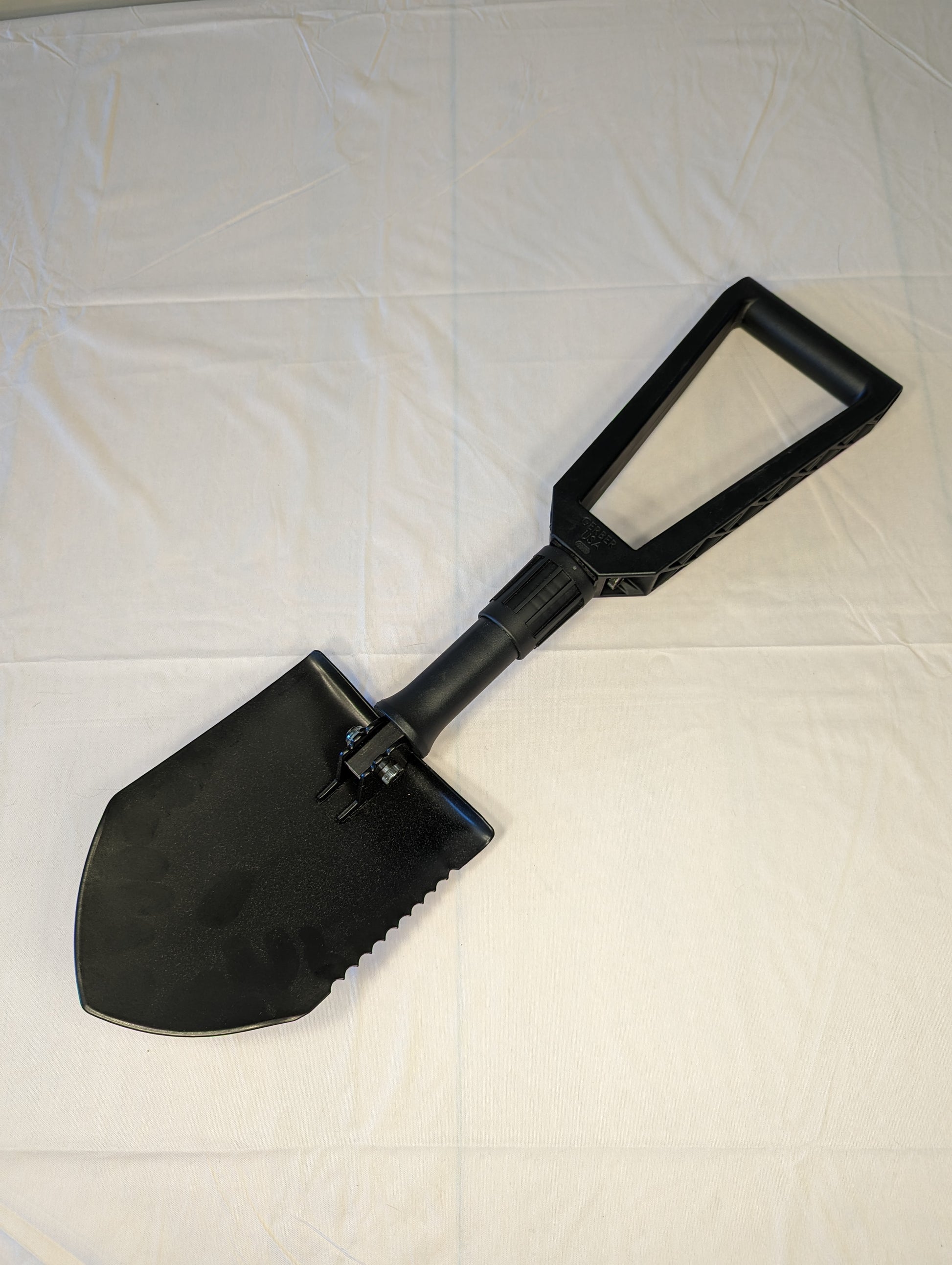 A black military surplus Gerber Entrenching Tool, partially folded to show the steel blade with a serrated edge and the black plastic D-handle, resting on a white background.