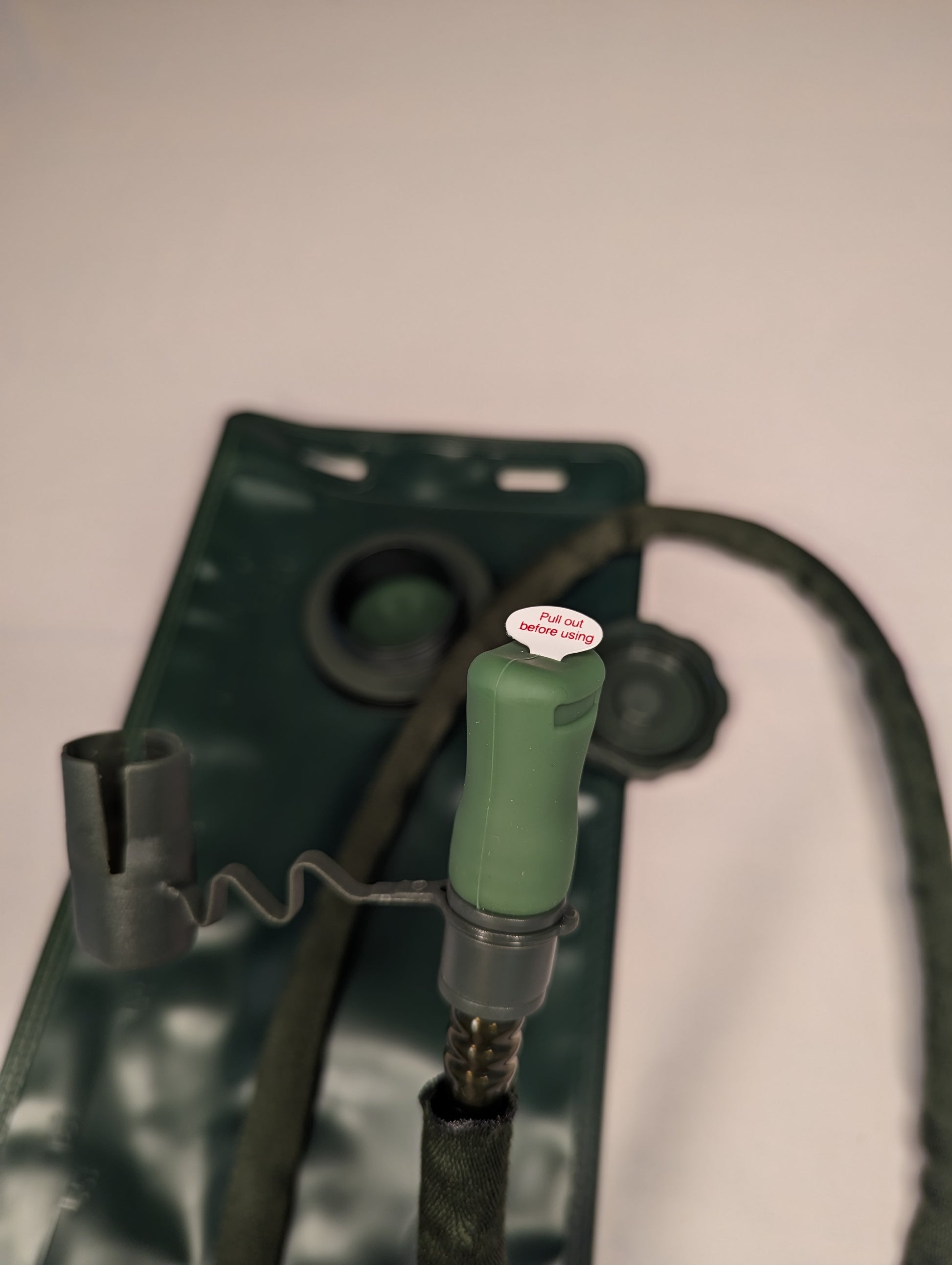 A close-up detail shot of the new dark green hydration bladder filler cap, showing the quick-connect hose attachment, coiled drinking tube, and bite valve.