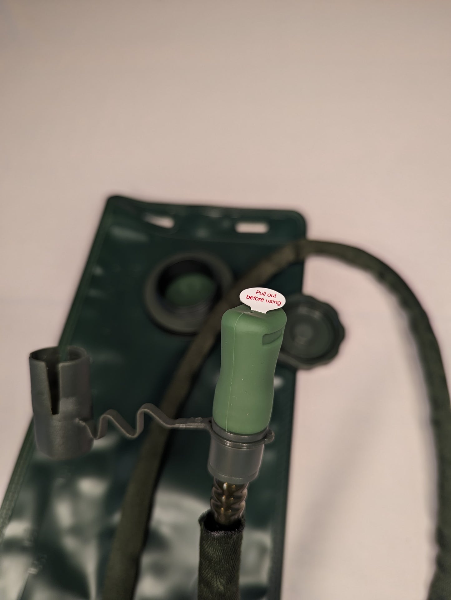 An extreme close-up of a new dark olive green hydration pack hose, showing the bite valve with a 'PULL OUT BEFORE DRINKING' safety tag, and the coiled insulated hose attachment.