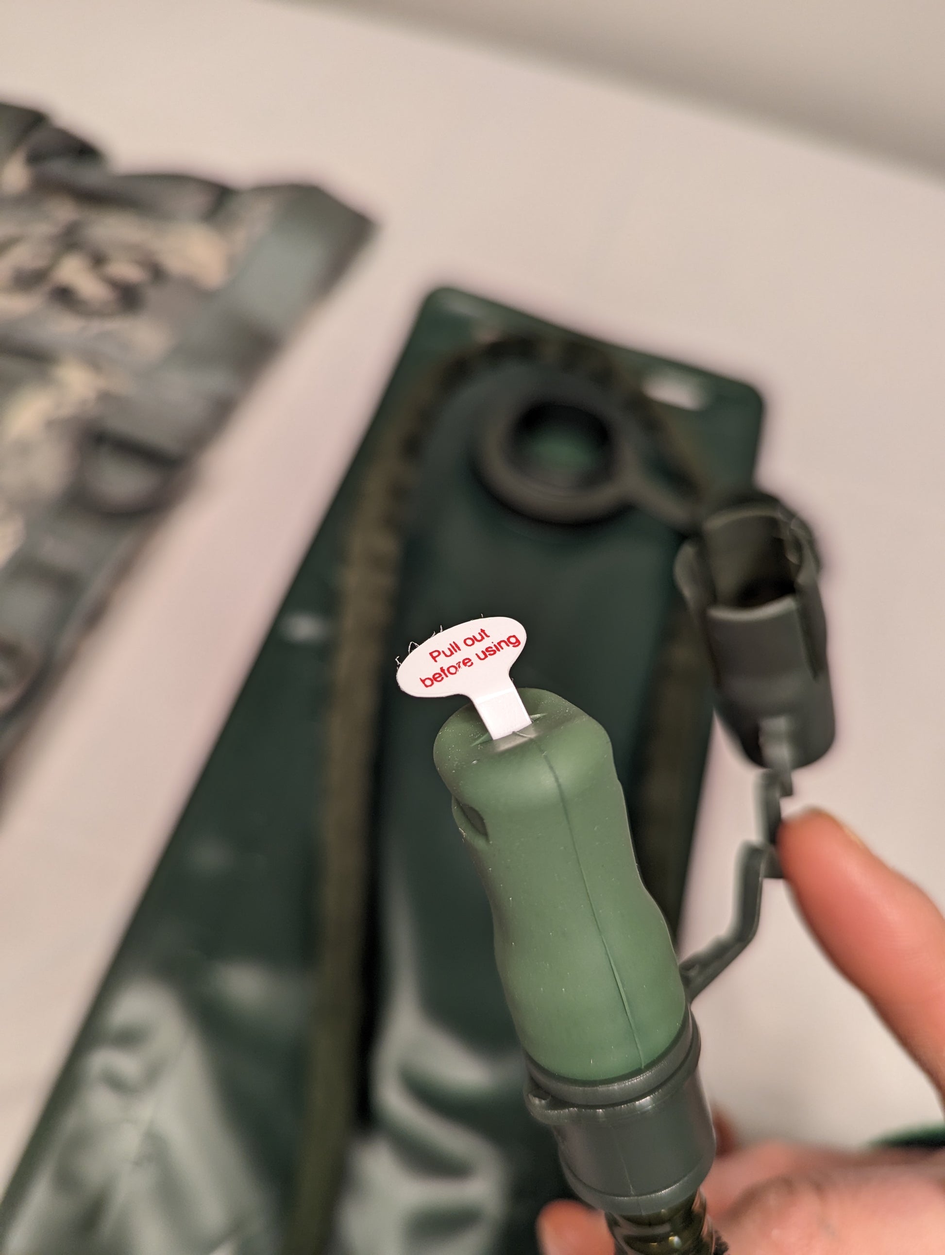 An extreme close-up of a hydration pack hose, showing the new green bite valve with a 'Pull out before drinking' safety tag, the quick-connect coupling, and the dark insulated hose, with a finger providing scale.