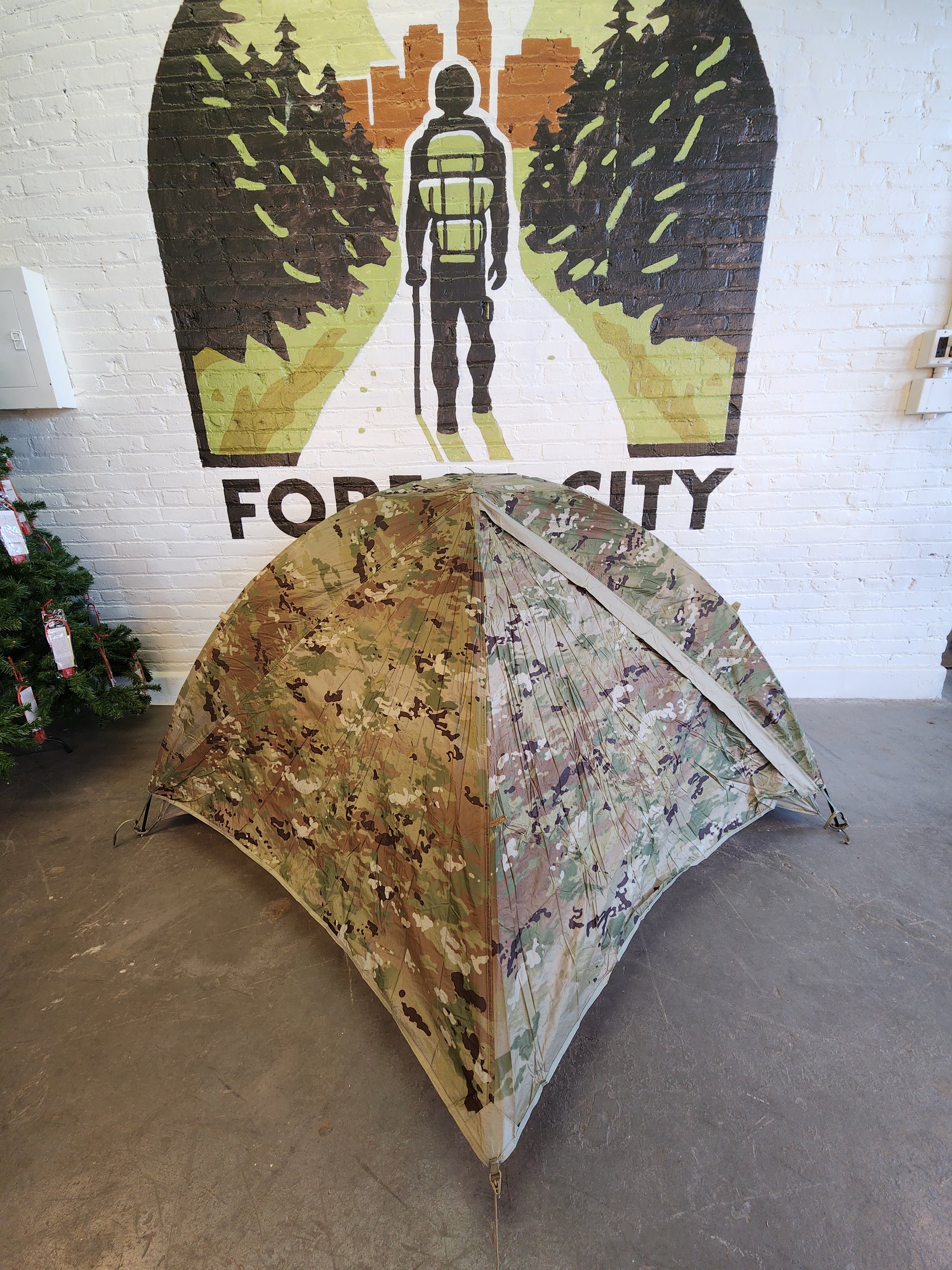 The inner mesh tent body of a Litefighter 1-Man Combat Shelter pitched indoors, featuring a tan waterproof floor and black mosquito netting, displayed in front of a painted Forest City Supply wall logo.