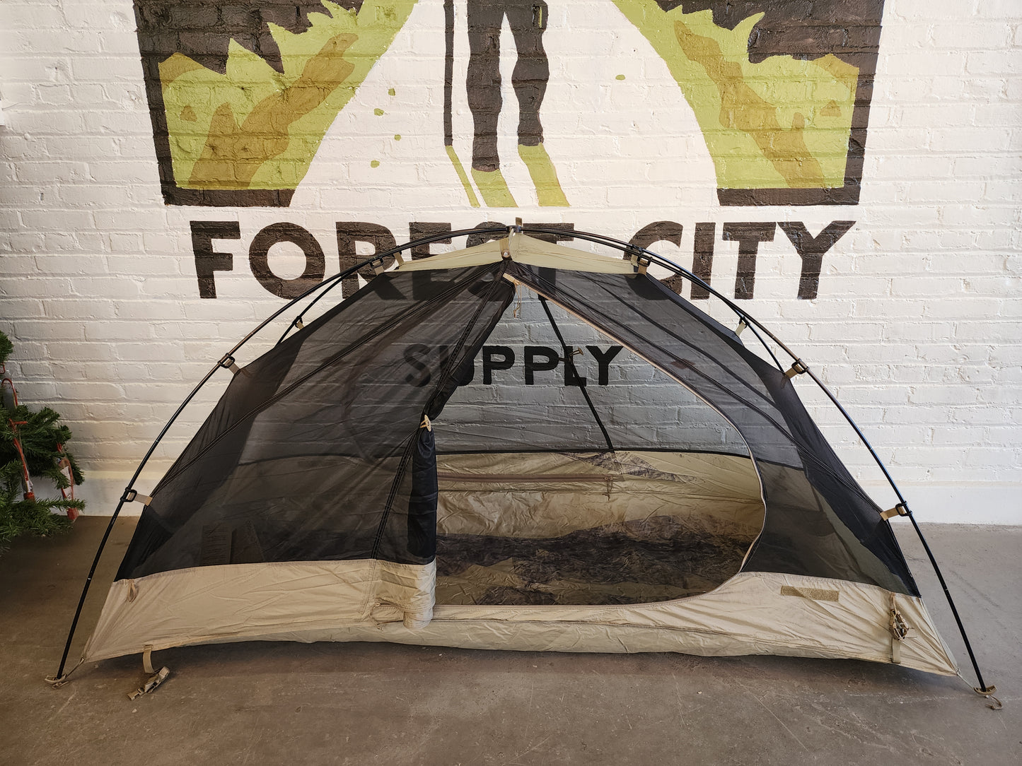 The inner mesh tent body of a Litefighter 1-Man Combat Shelter pitched indoors, featuring a tan waterproof floor and black mosquito netting, displayed in front of a painted Forest City Supply wall logo.