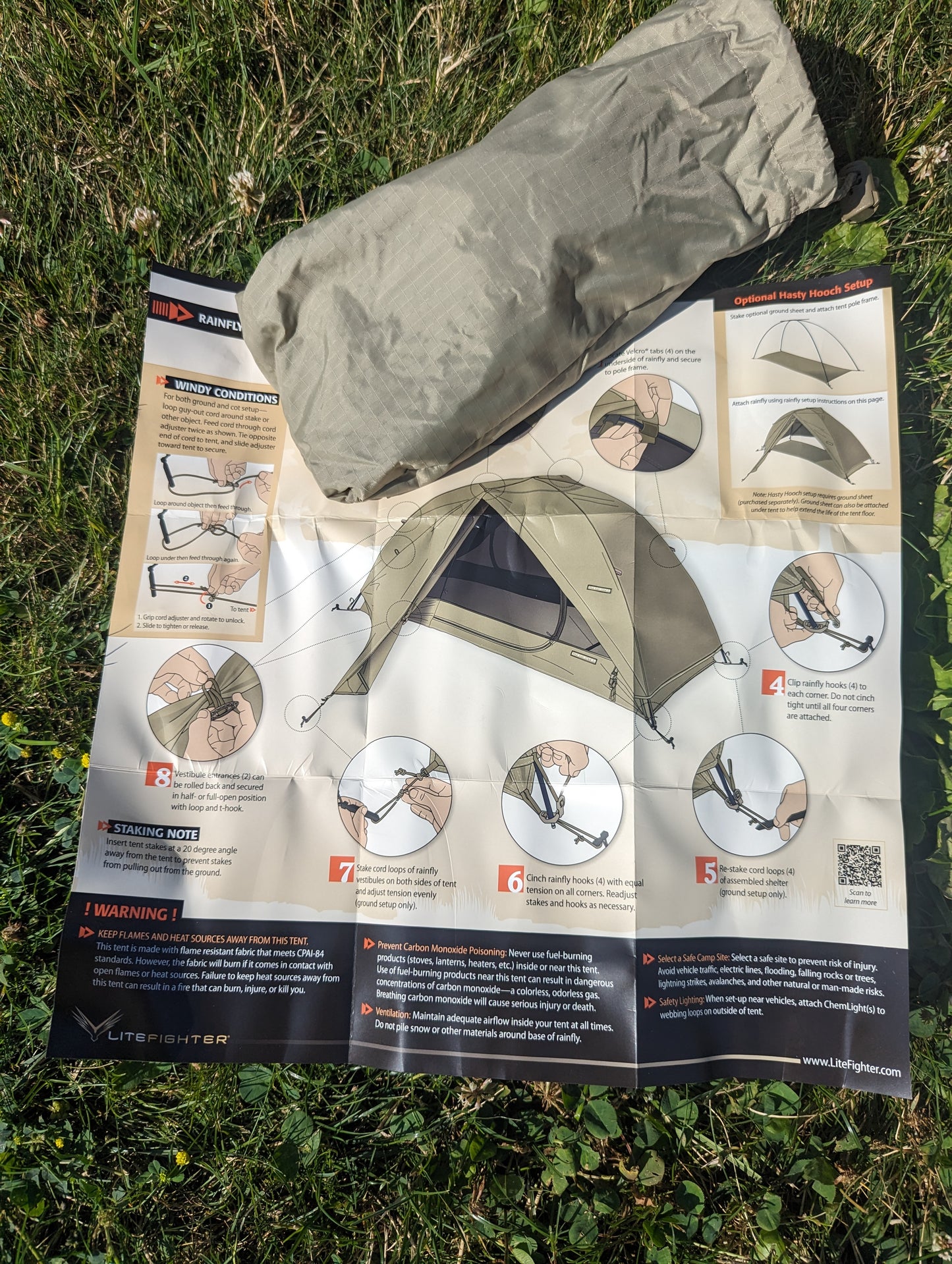 An overhead view of the U.S. Military Litefighter 1-Man Combat Shelter instruction manual laid on green grass, featuring step-by-step setup diagrams and a rolled-up tan tent component.