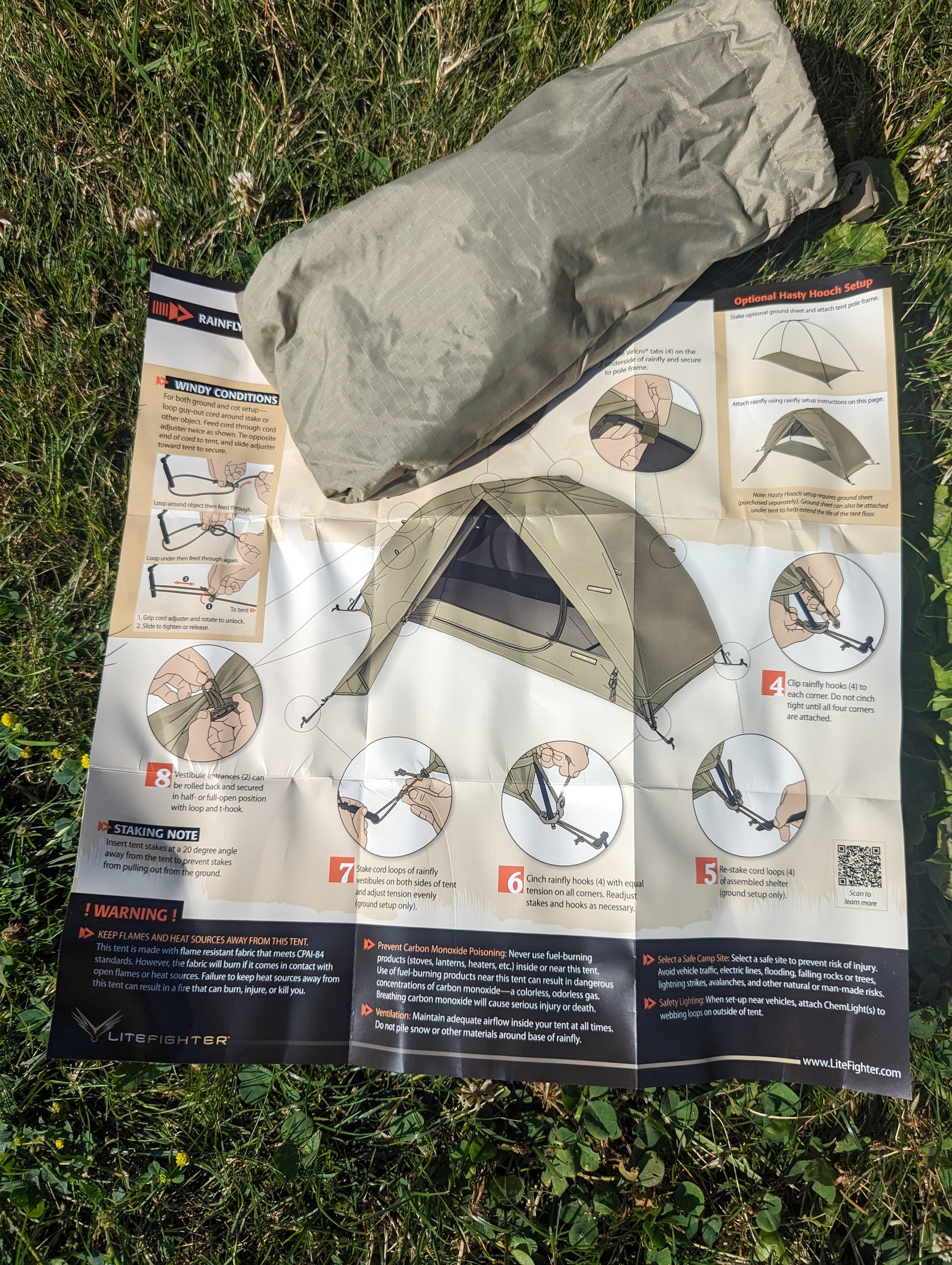 An overhead view of the U.S. Military Litefighter 1-Man Combat Shelter instruction manual laid on green grass, featuring step-by-step setup diagrams and a rolled-up tan tent component.