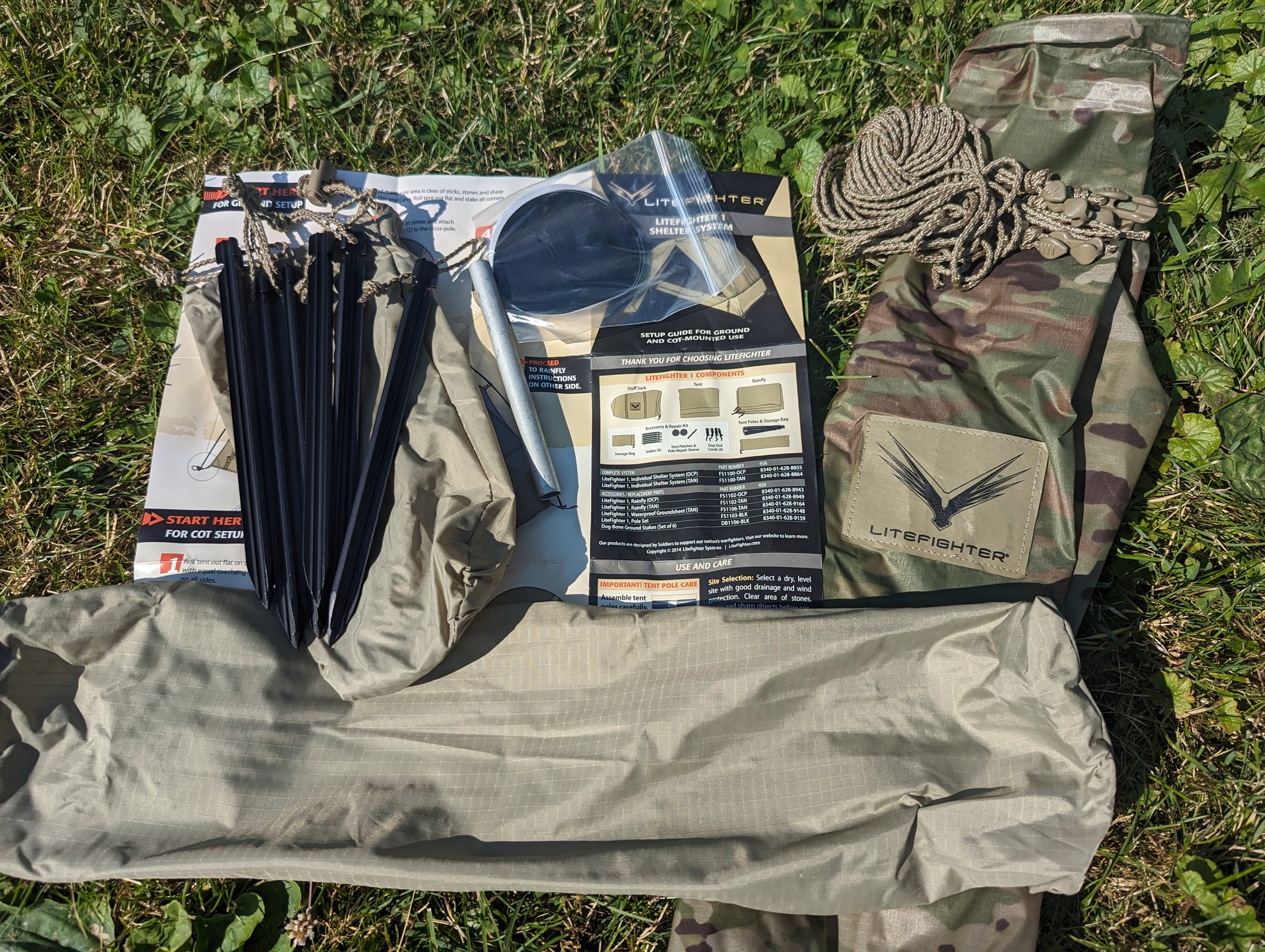An overhead flat lay photo of the entire Litefighter 1-Man Combat Shelter kit, laid out on grass. The kit includes the OCP/Multicam storage bag, tent poles, instruction manual, cord, and the main tent fabric.