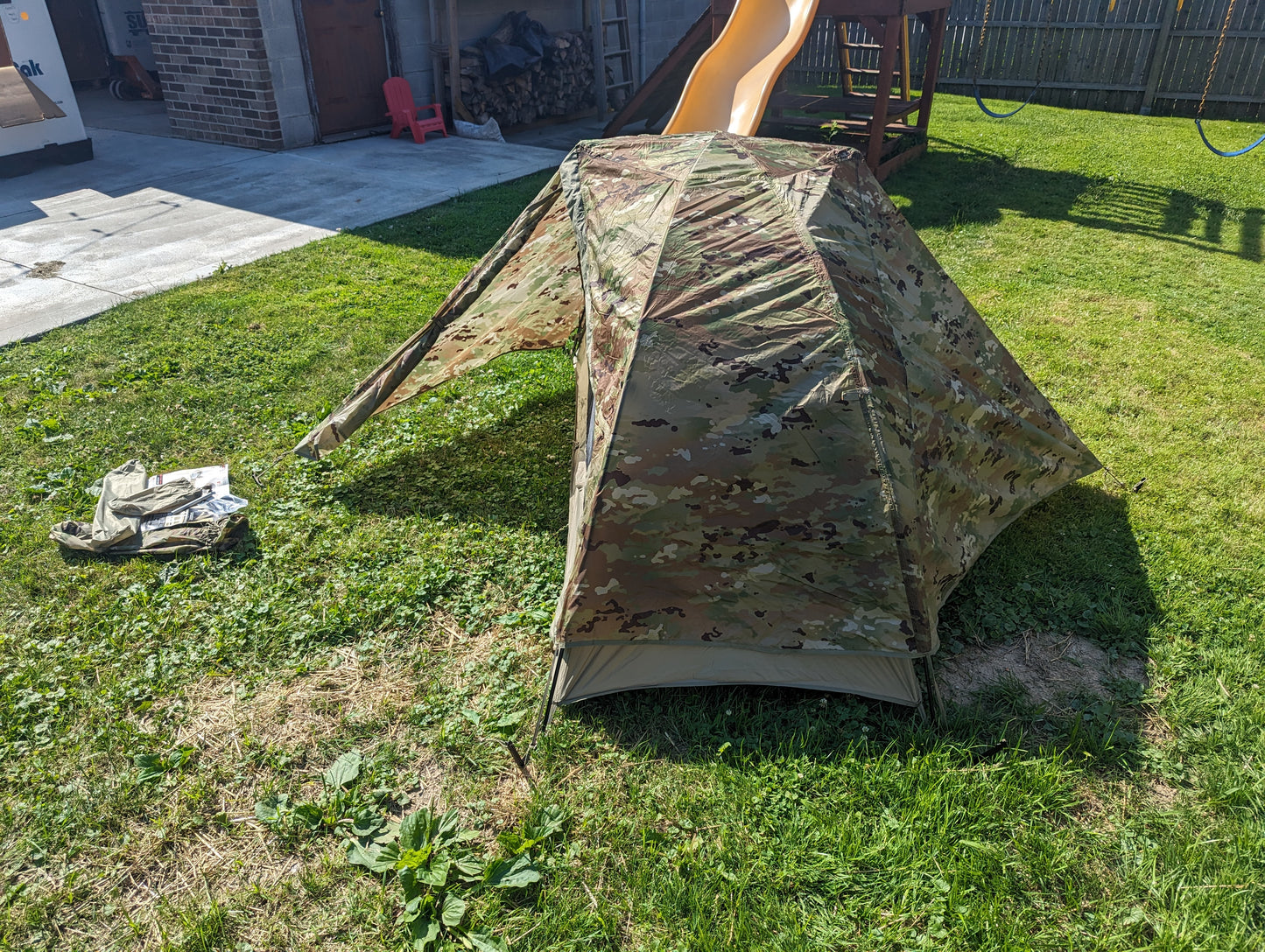 A fully pitched U.S. Military Litefighter 1-Man Combat Shelter in OCP/Multicam camouflage, set up on a grassy lawn with its front vestibule open, showcasing the low-profile tent and the domestic backyard setting.