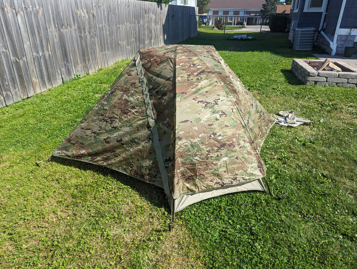 A fully pitched U.S. Military Litefighter 1-Man Combat Shelter in OCP/Multicam camouflage, set up on a grassy lawn with a wooden fence and a fire pit visible in the background.