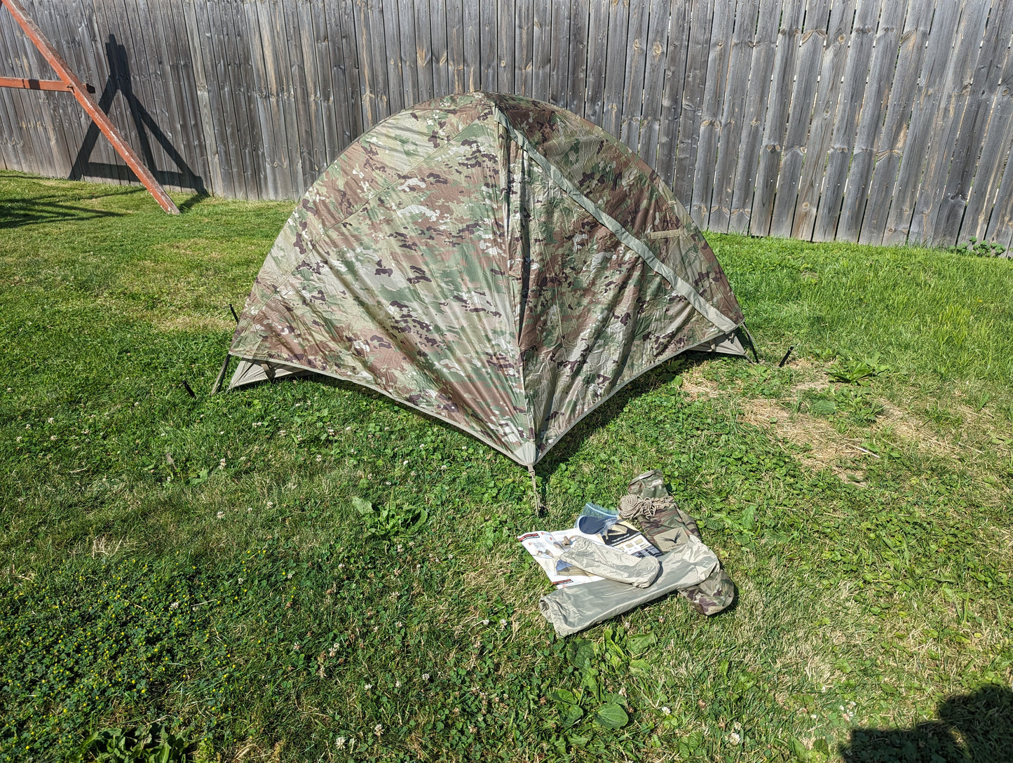 A low-profile U.S. Military Litefighter 1-Man Combat Shelter in OCP/Multicam camouflage, fully pitched on a grassy lawn, with field gear and a map laid out in the foreground.