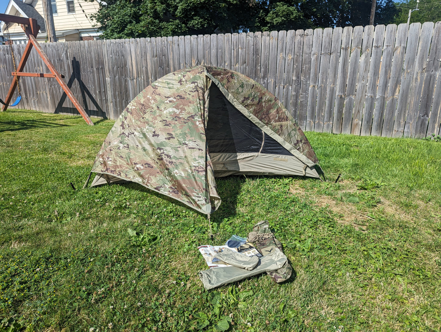 A fully pitched U.S. Military Litefighter 1-Man Combat Shelter in OCP/Multicam camouflage, set up on a grassy lawn with the door open and the storage bag resting in the foreground.
