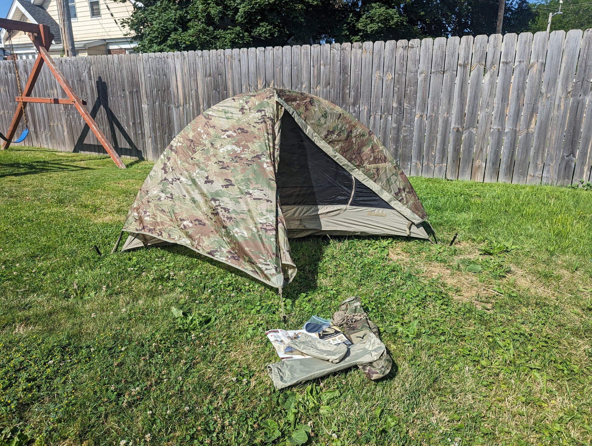 A fully pitched U.S. Military Litefighter 1-Man Combat Shelter in OCP/Multicam camouflage, set up on a grassy lawn with the door open and the storage bag resting in the foreground.