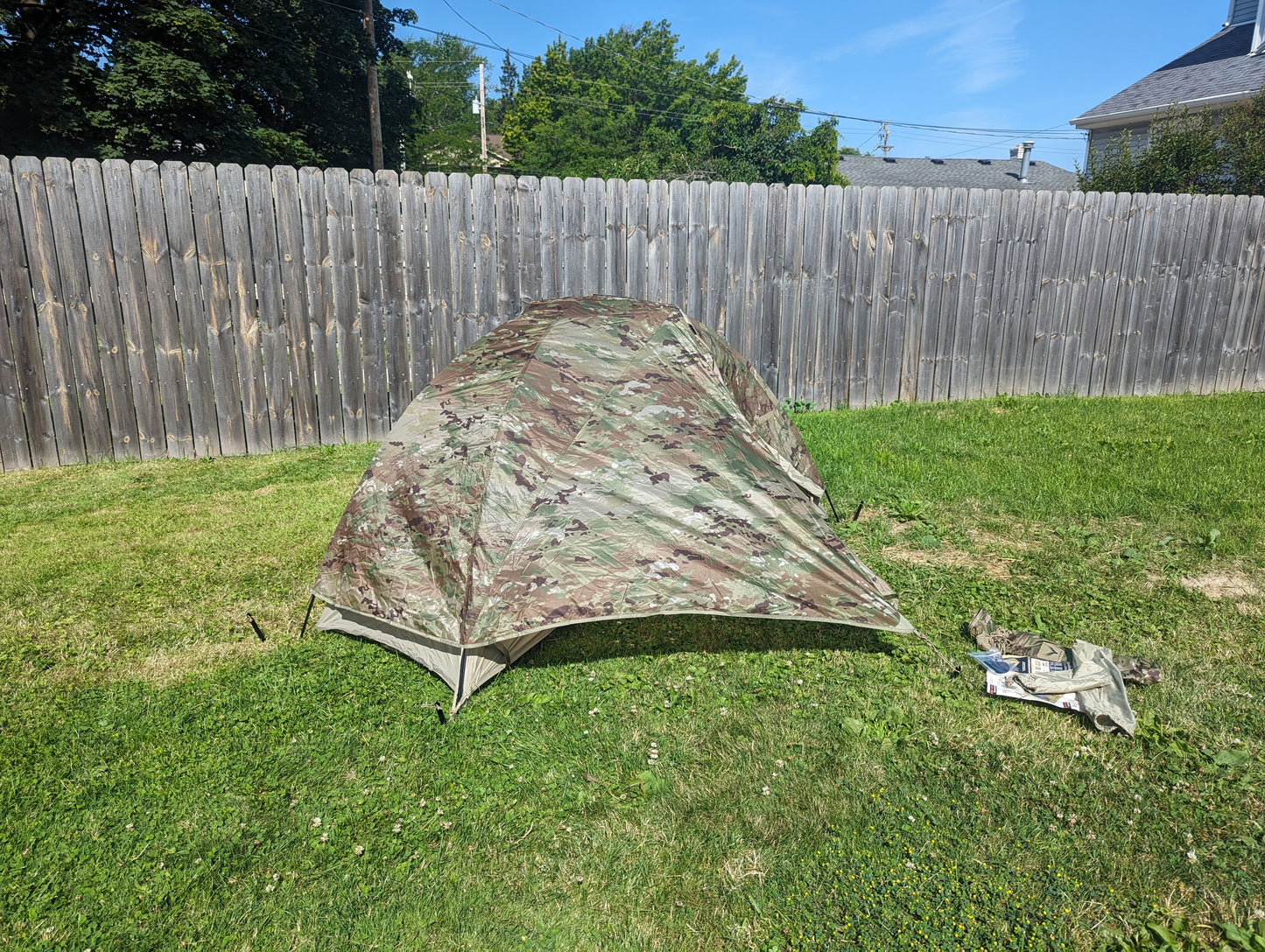 A U.S. Military Litefighter 1-Man Combat Shelter in OCP/Multicam camouflage, fully pitched on a green lawn with a wooden fence background, and a small storage bag resting nearby.