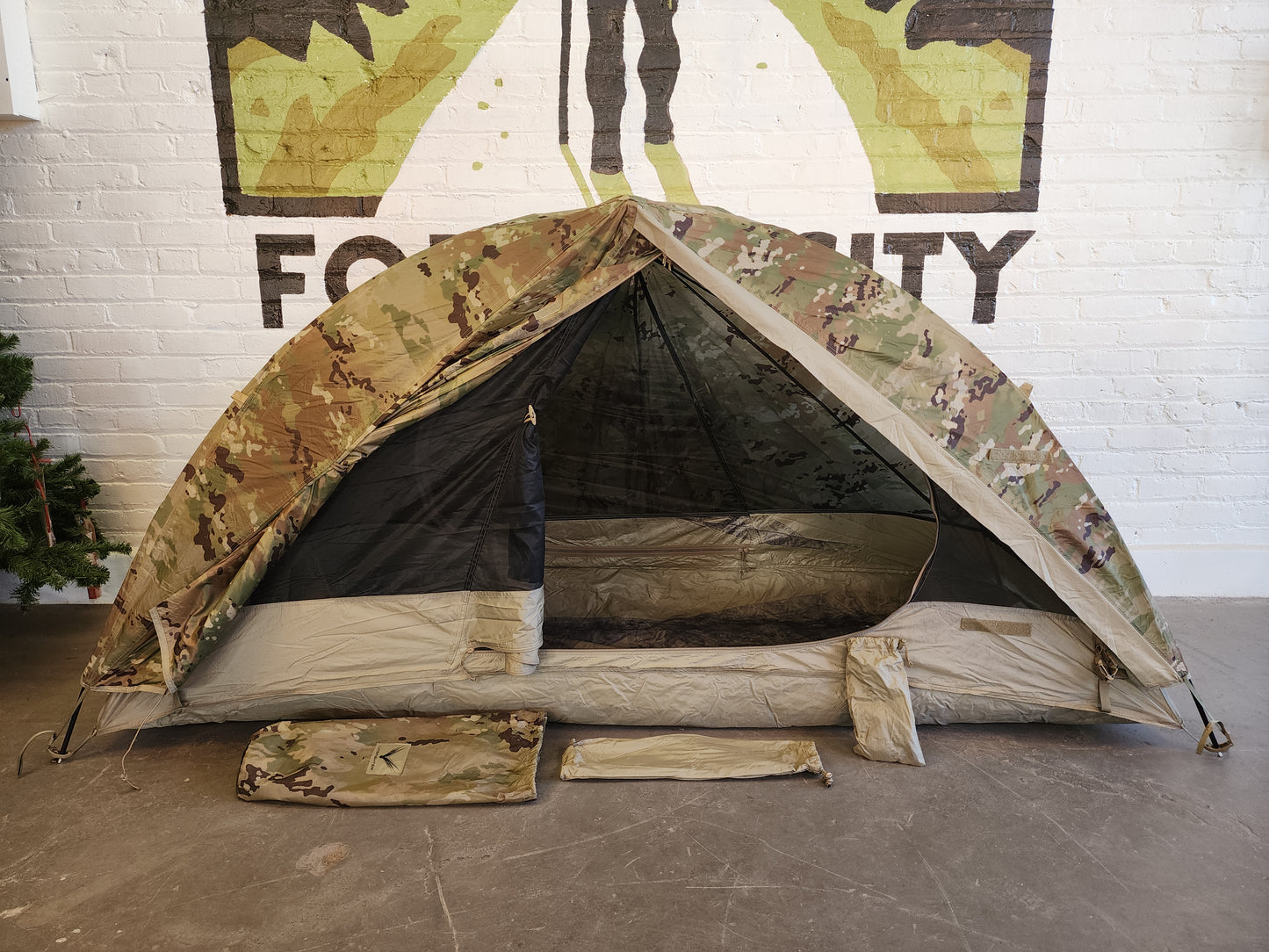 A Litefighter 1-Man Combat Shelter in OCP/Multicam camouflage pitched on a concrete floor, with the rainfly door rolled back to reveal the inner mesh tent, and storage bags displayed in front.