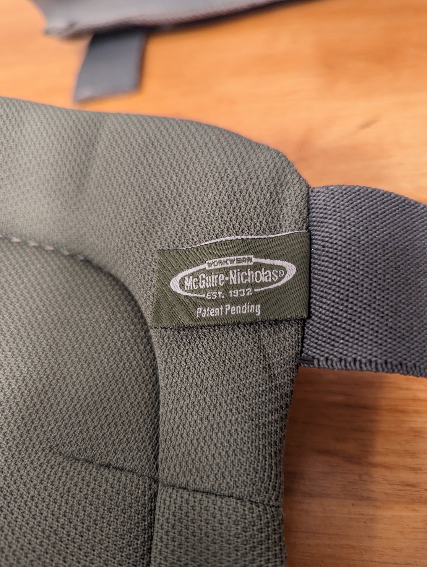 A close-up photograph of the McGuire-Nicholas brand label sewn onto a piece of dark olive green padded tactical gear, displaying the text 'Patent Pending' and the company's establishment date.