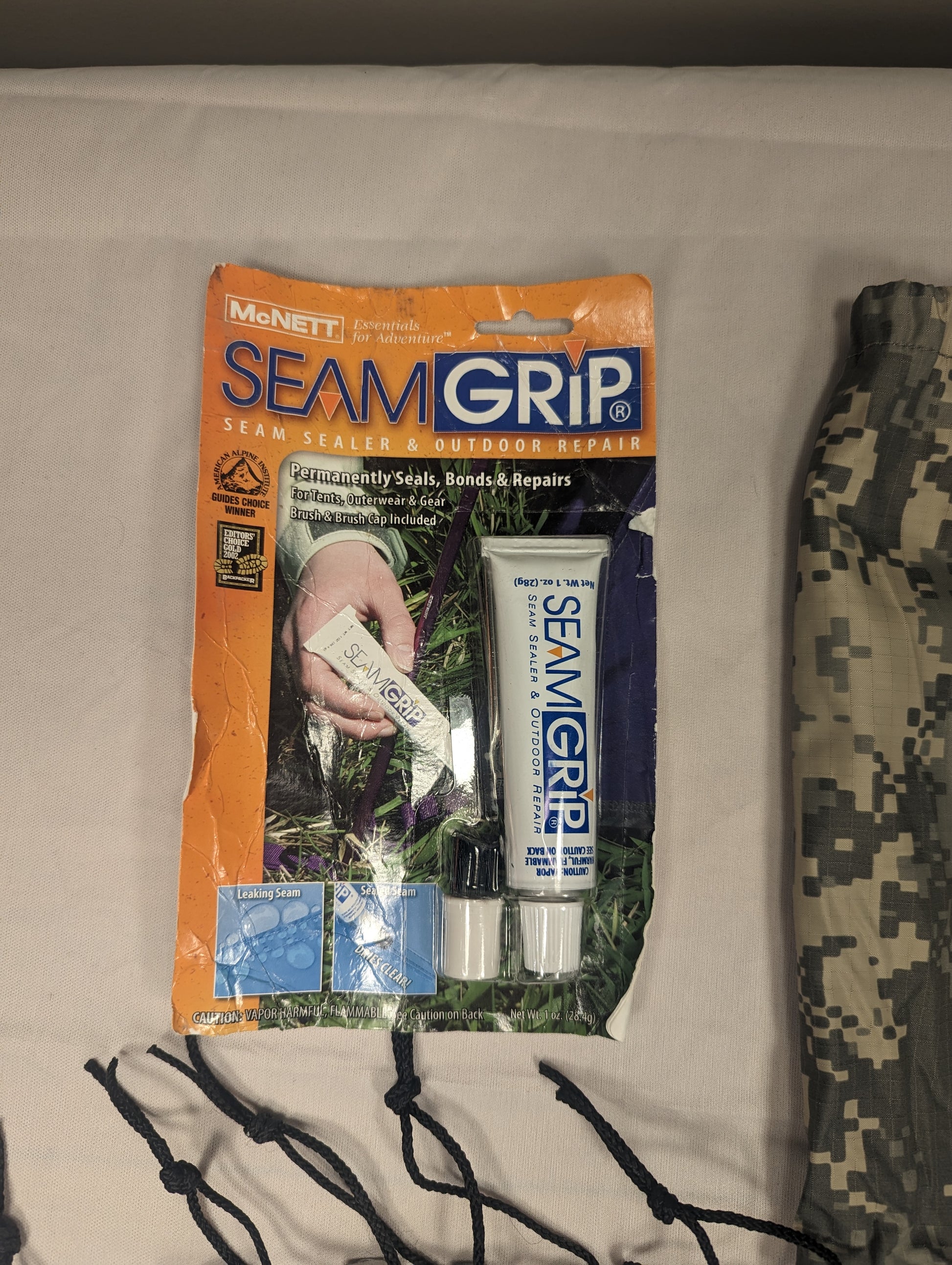 A packaged tube of McNett Seam Grip polyurethane sealant, displayed next to a rolled-up ACU digital camouflage tent component and black repair cords, part of the ICS Repair Kit.