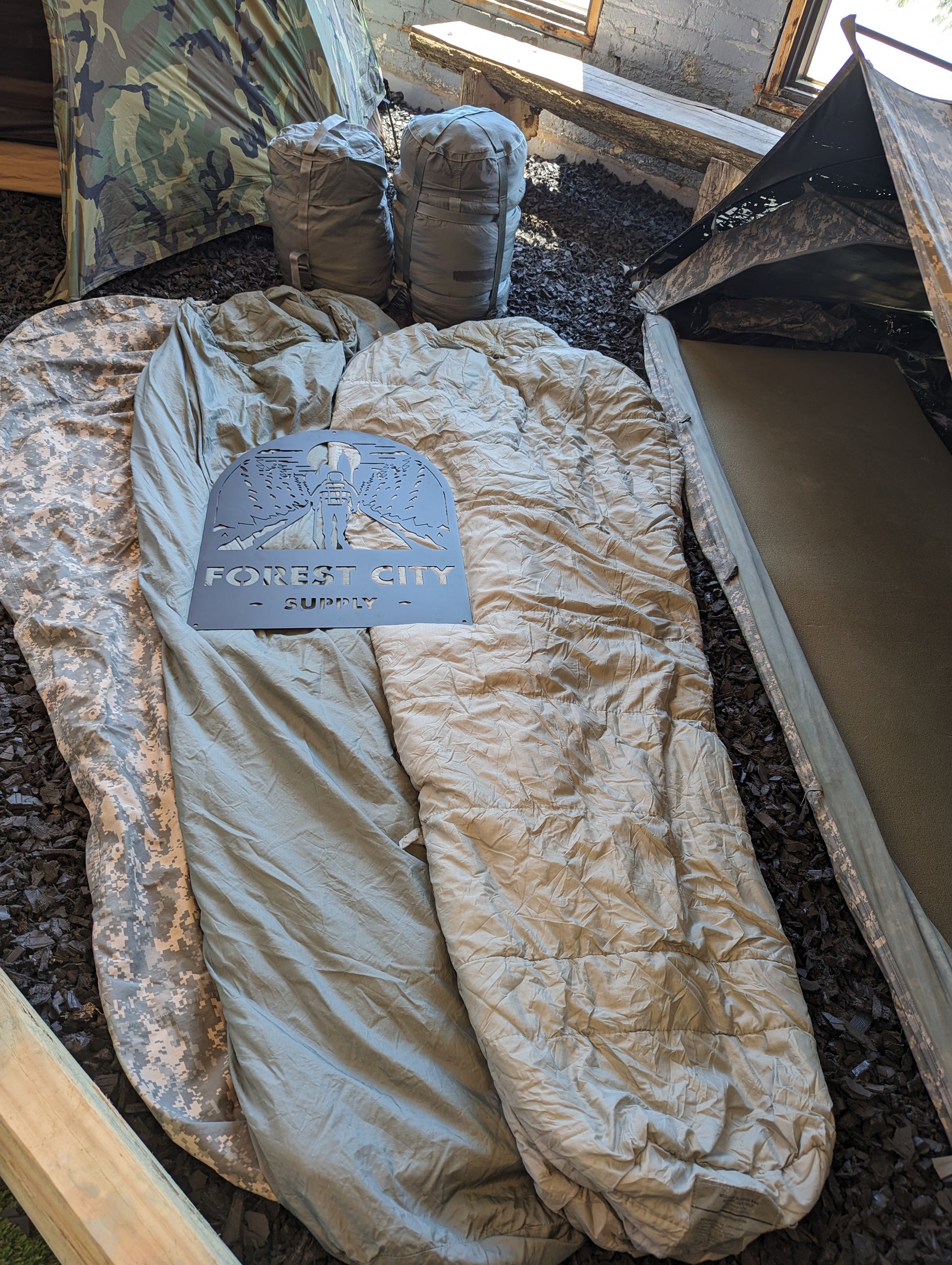 A light olive green U.S. Military Modular Sleep System Patrol Sleeping Bag laid open in a field environment, with the "FOREST CITY SUPPLY" logo stencil resting on the fabric.