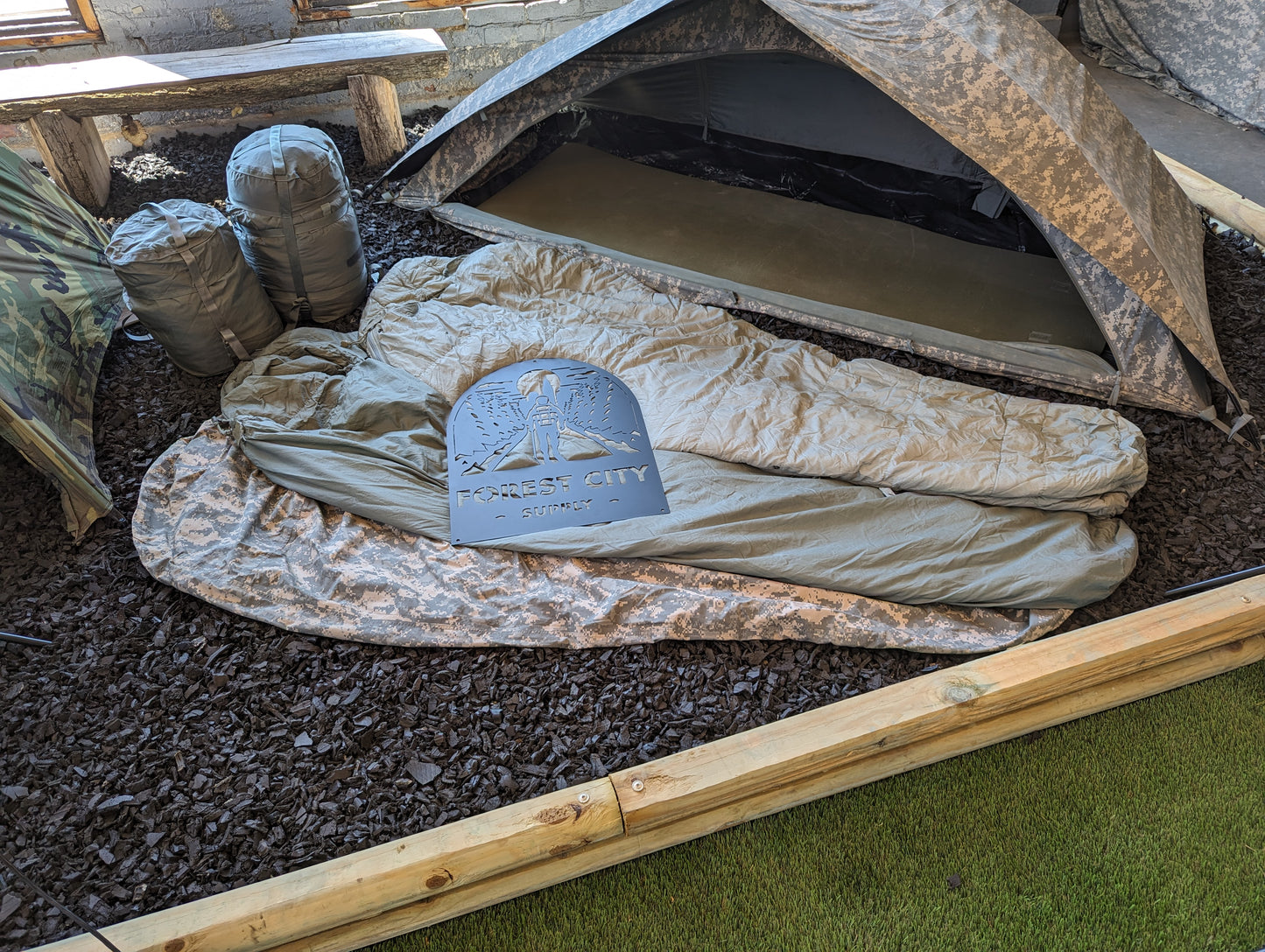 A light olive green U.S. Military Modular Sleeping Bag laid open in a tactical setting, with the "FOREST CITY SUPPLY" logo stencil resting on the fabric, and a camouflage shelter pitched behind it.