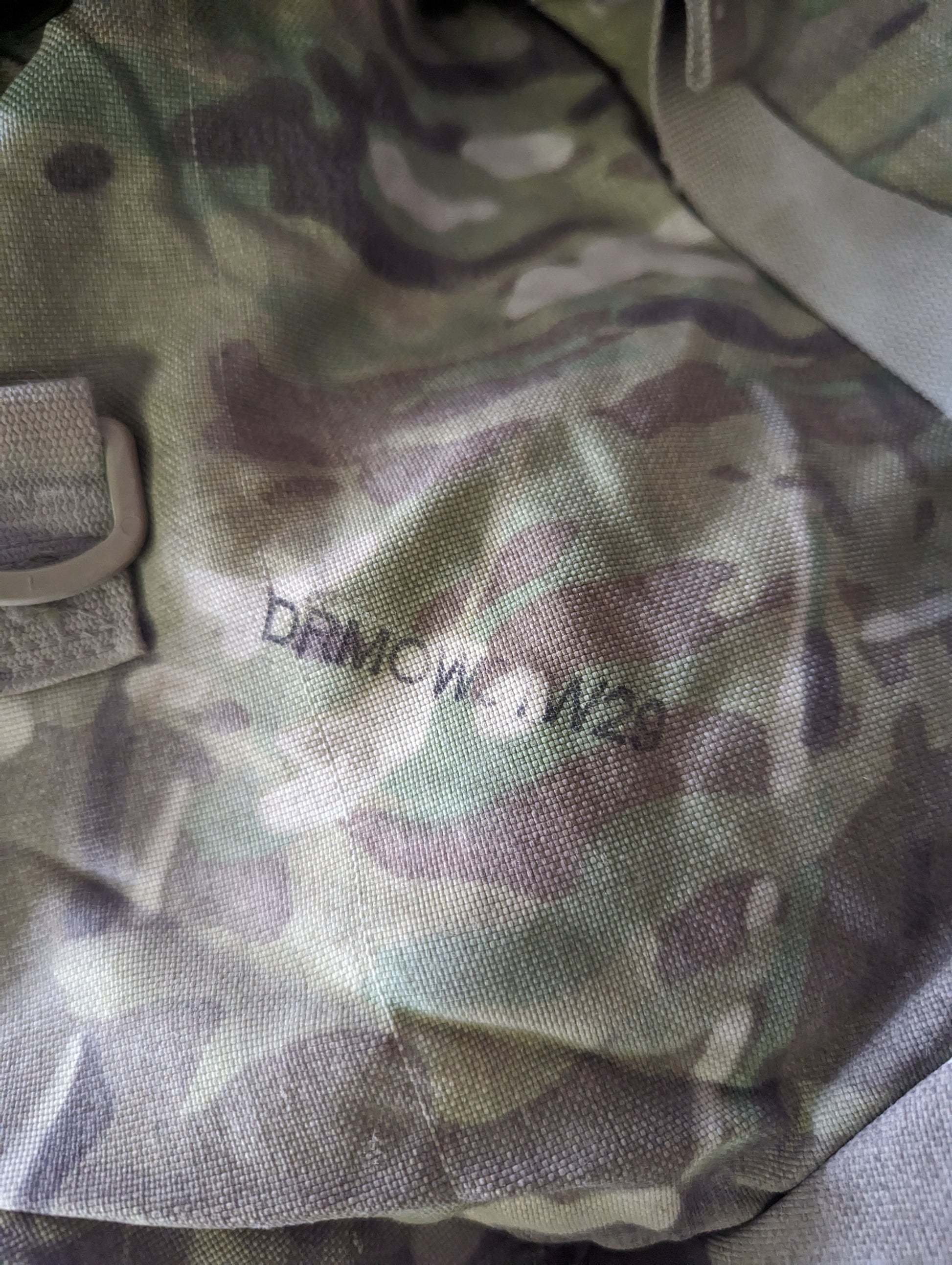 A close-up photograph of OCP/Multicam camouflage fabric, highlighting the weave of the material and a stenciled military identification marking that reads 'DRMCWG 1125'.