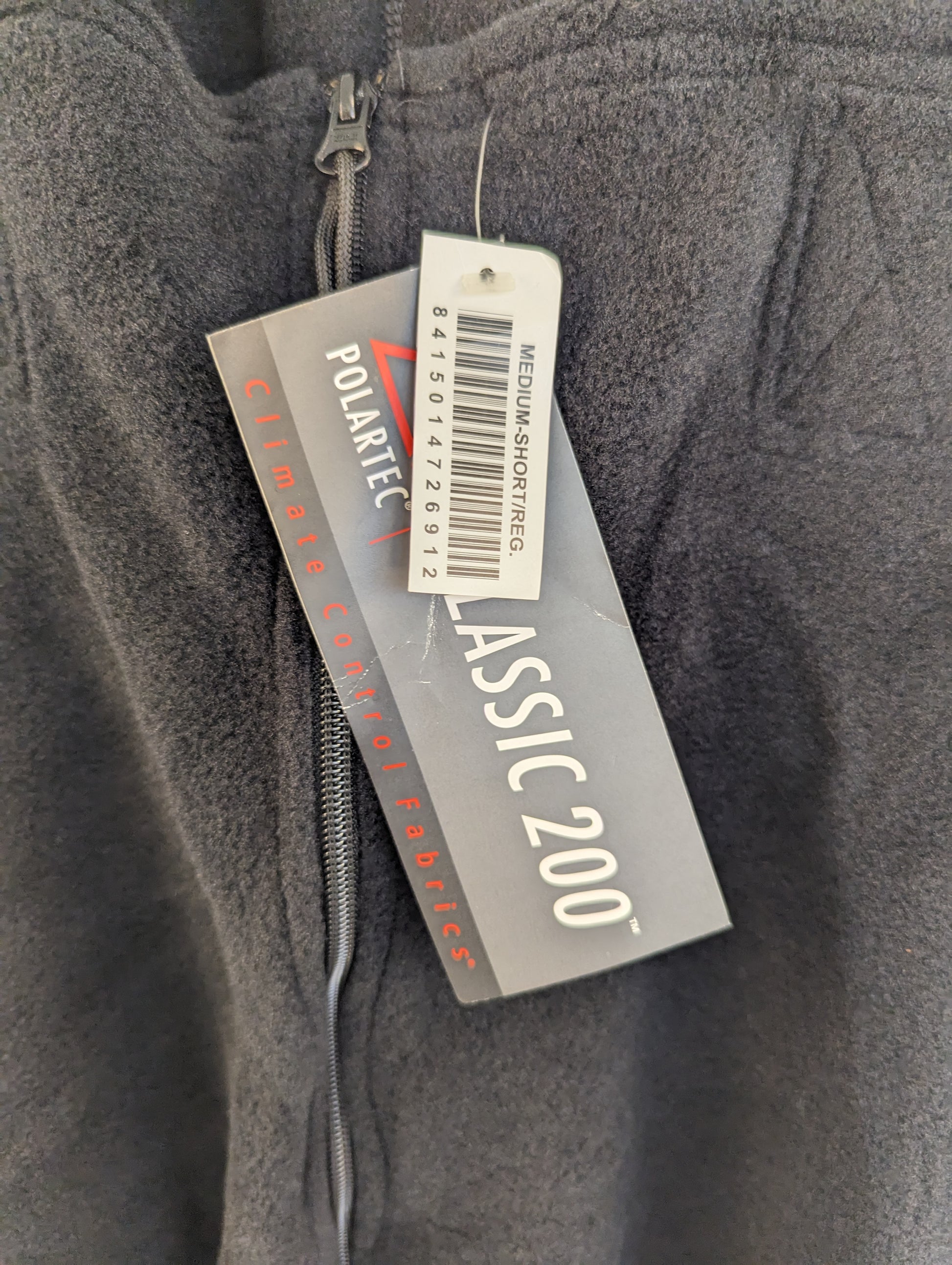 A close-up of the Polartec Classic 200 hang tag attached to the zipper of black fleece cold weather overalls, displaying the size 'MEDIUM-SHORT/REG' and barcode.