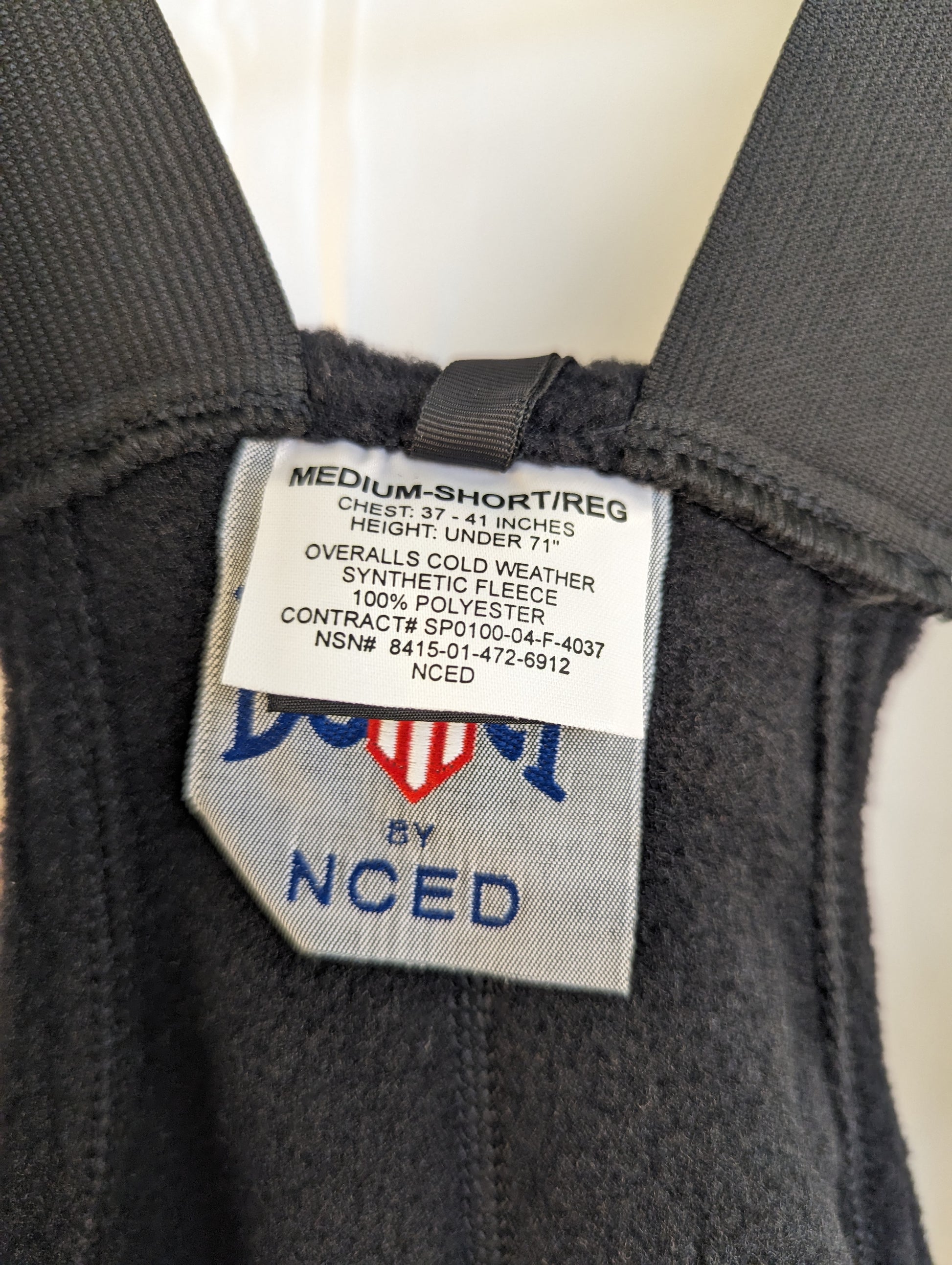 A close-up photograph of the issue tag sewn into the black Polartec fleece overalls, displaying the size 'MEDIUM-SHORT/REG', material details, and the National Stock Number (NSN).